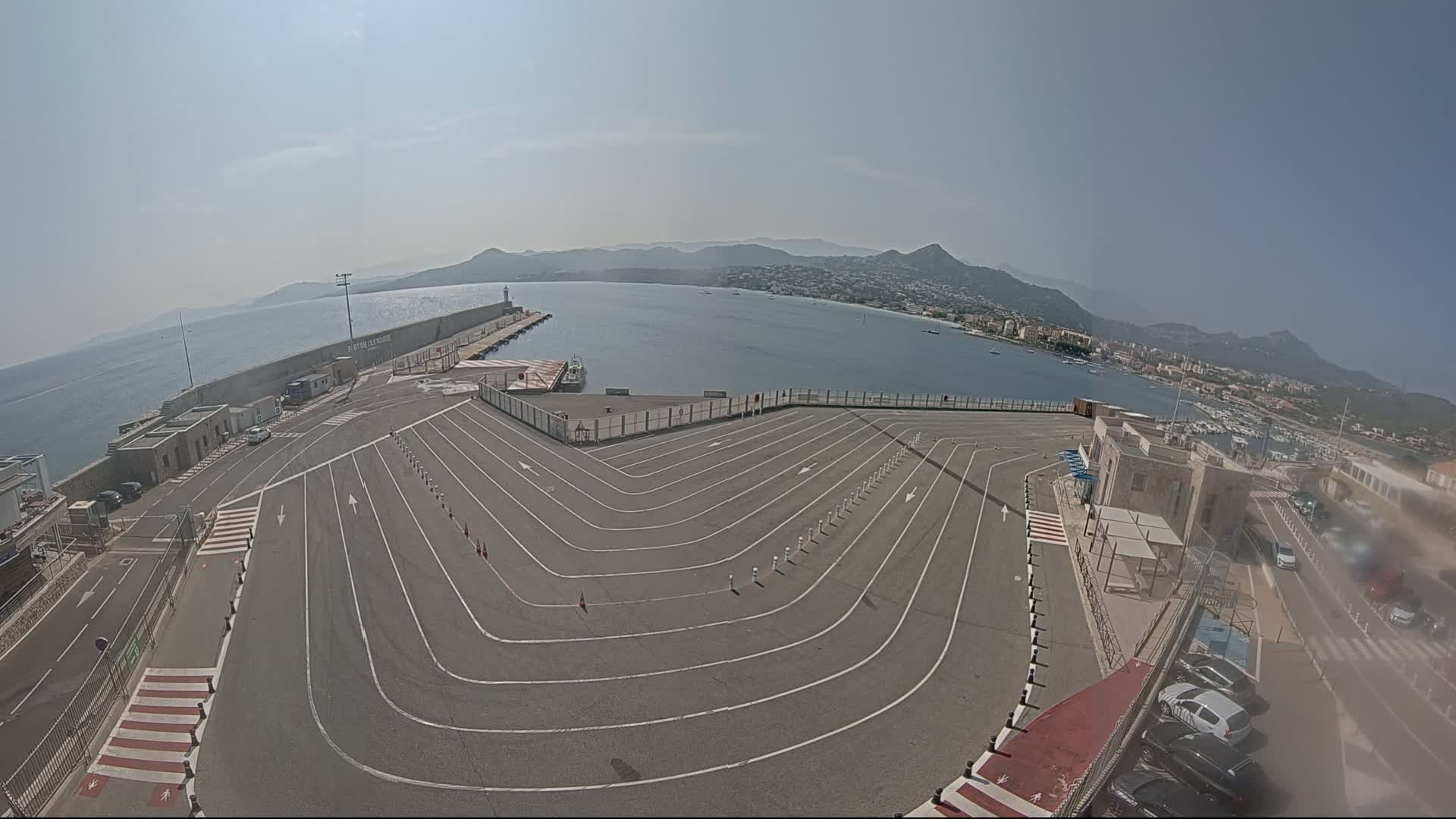 An empty, curved roadway at a harbor overlooks a calm body of water and distant hills under a clear, sunny sky.