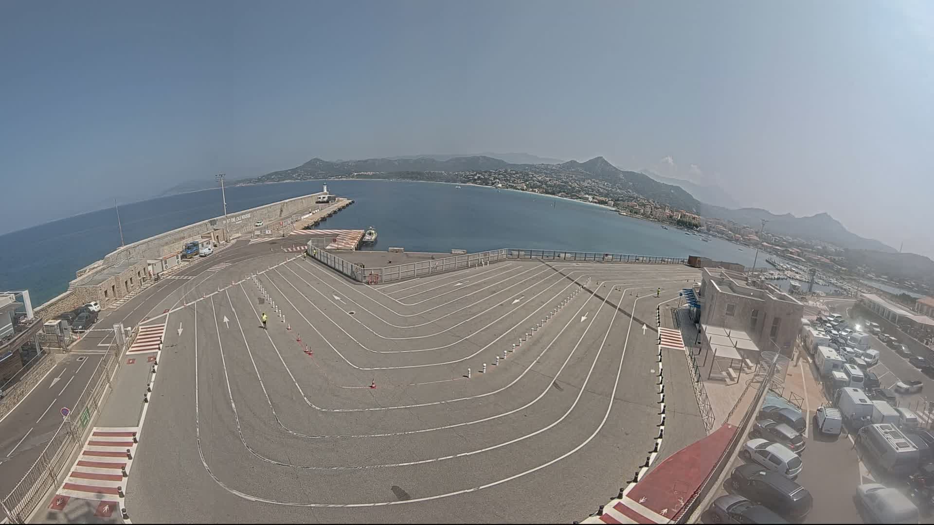A paved area with marked lanes curves around a harbor, overlooking a calm sea and distant mountains under a clear, sunny sky.