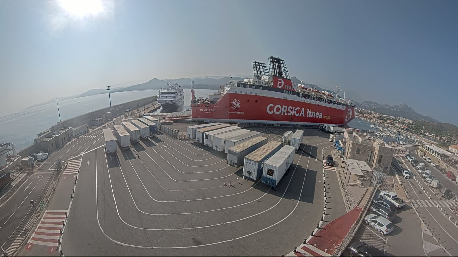 A large red ferry is docked at a port on a sunny day, with several semi-trailer trucks parked nearby and another smaller ship visible in the distance.
