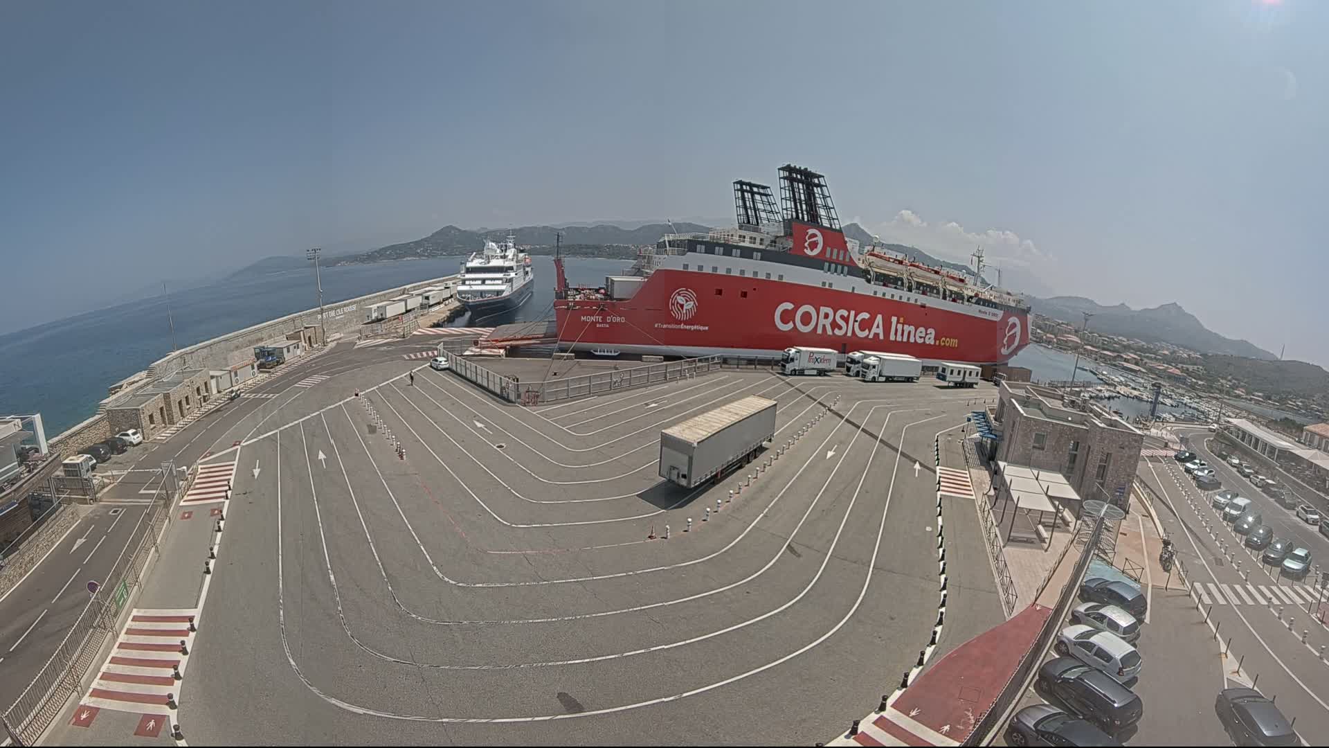 A large red ferry is docked at a port on a sunny day, with another smaller ship nearby and several vehicles parked in the surrounding area.