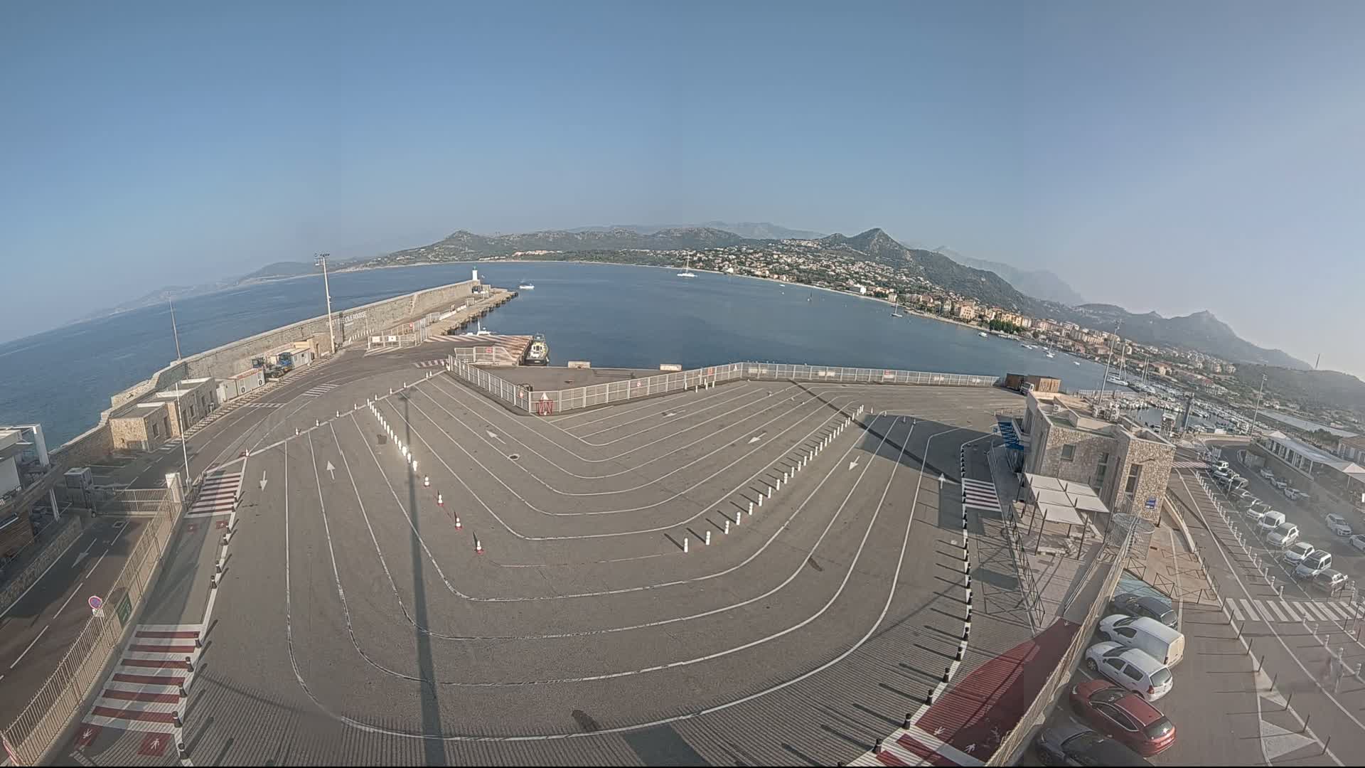 An empty, marked parking lot at a harbor overlooks a calm bay surrounded by mountains under a clear blue sky.
