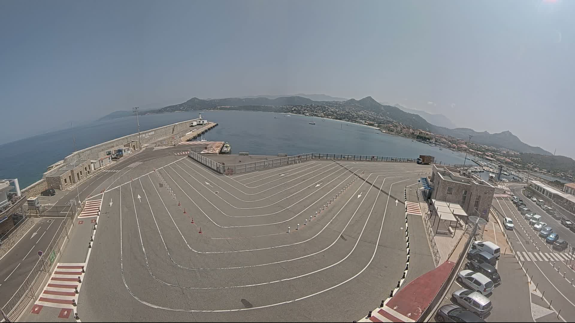 A paved area with marked lanes leading to a harbor, overlooking a calm body of water and distant mountains under a clear, sunny sky.