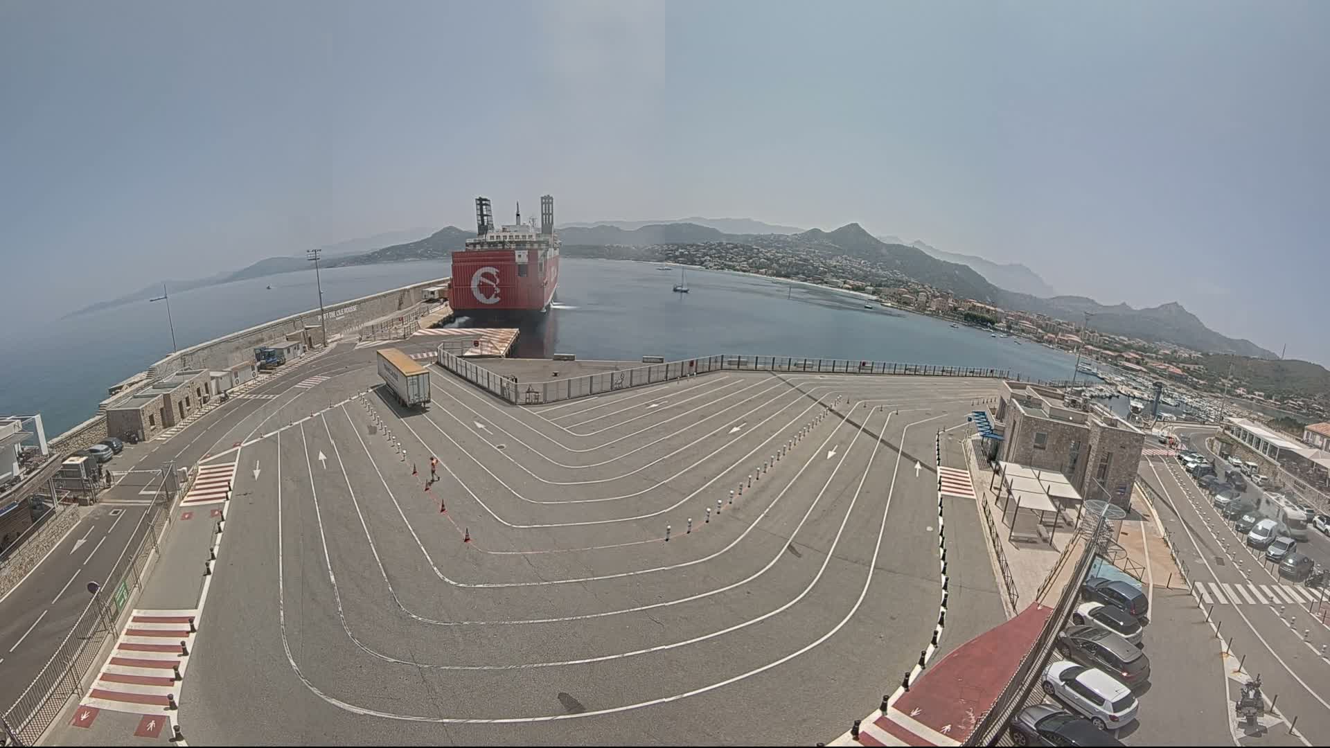 A large red ship is docked at a port on a sunny day, with a paved area marked with lines for vehicle maneuvering, and a car park is visible to the right of the image.