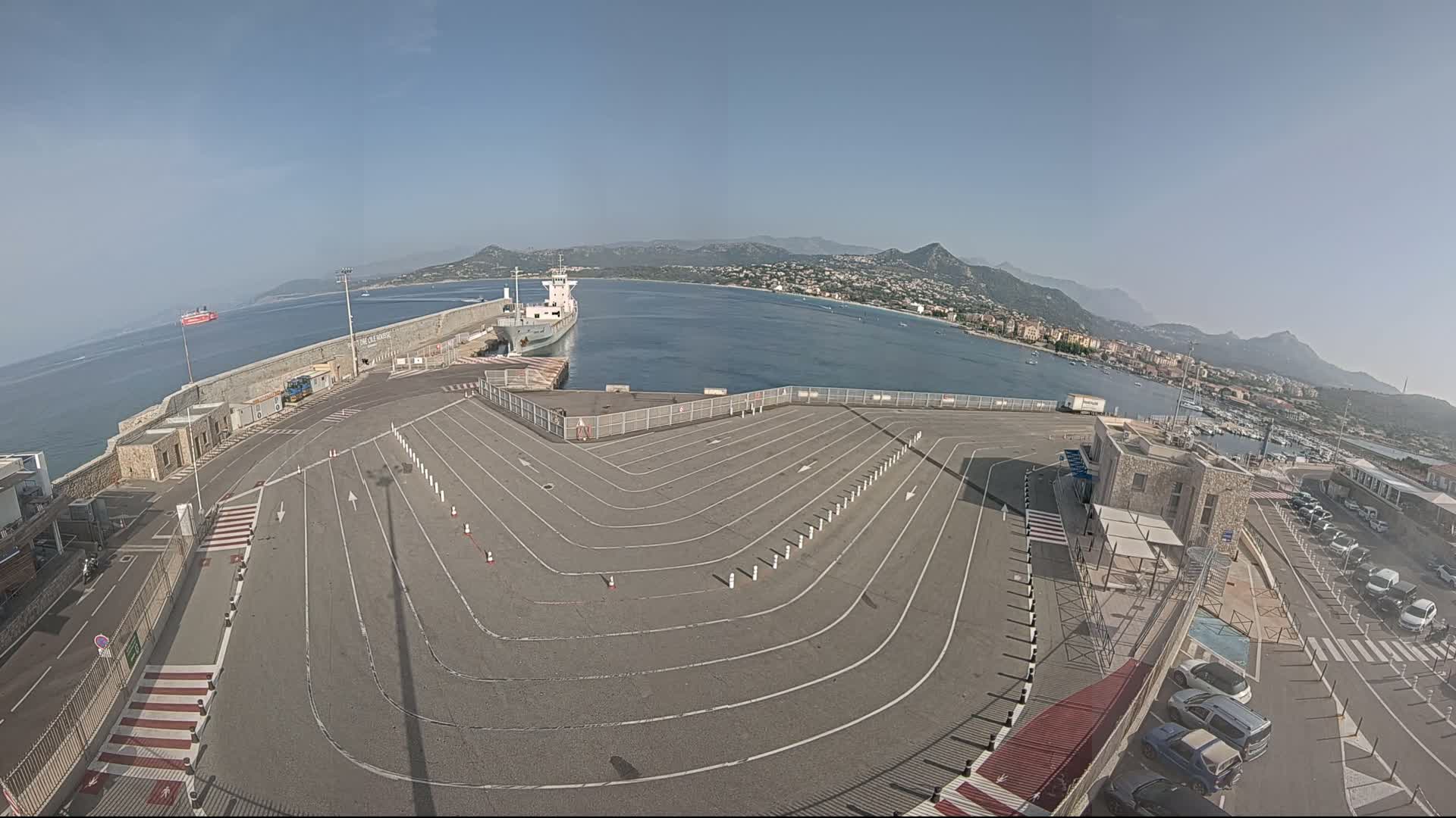 A large, empty vehicle staging area at a ferry terminal overlooks a calm bay with a ship docked and mountains in the background on a clear, sunny day.