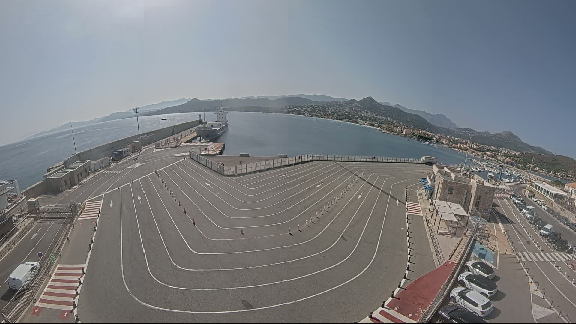 A large, empty ferry terminal with marked lanes curves toward a calm body of water, where a ship is docked, under a clear, sunny sky, with mountains visible in the distance.