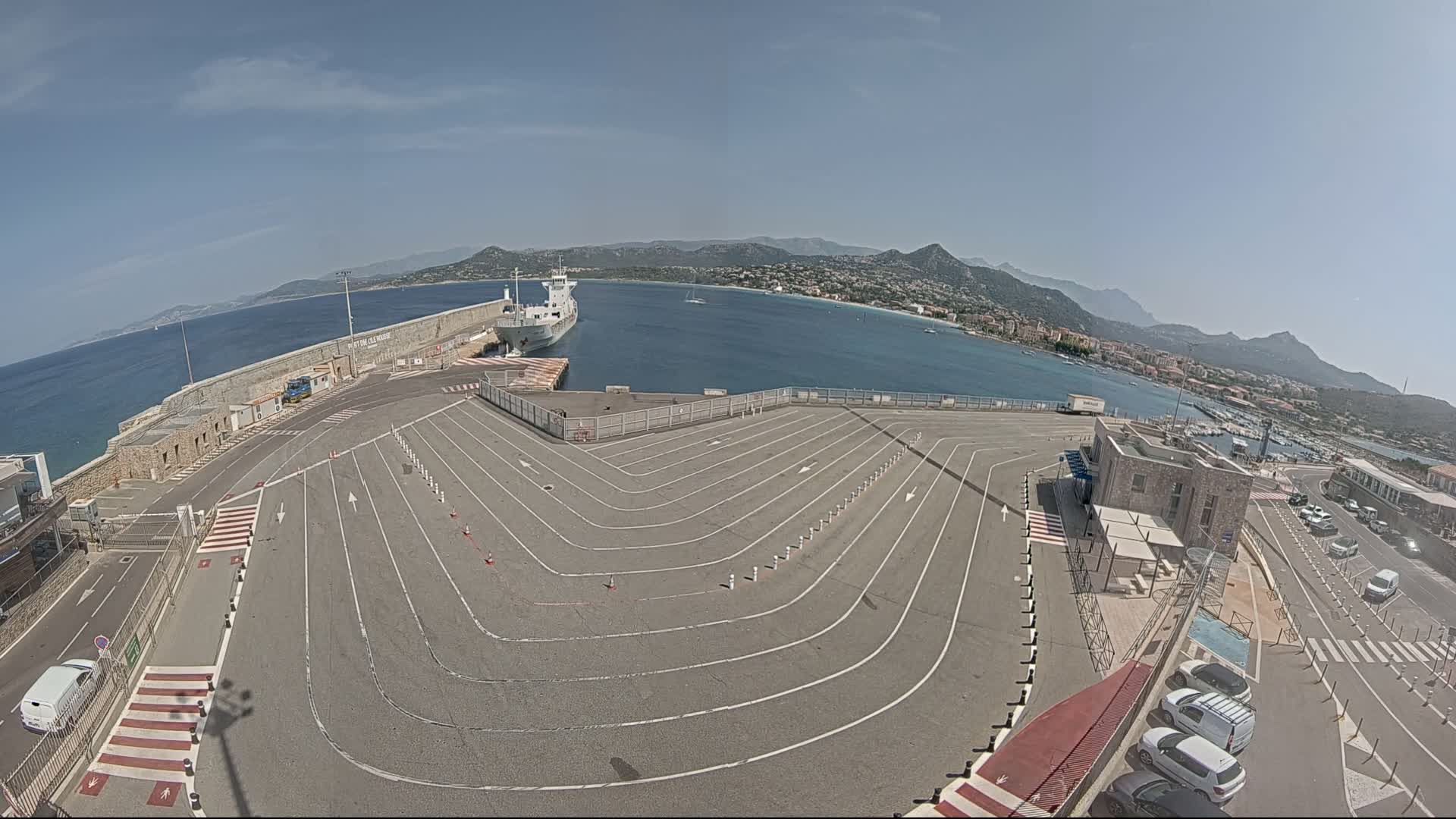 A large, empty paved area with marked lanes curves toward a docked ship and a body of water under sunny skies, backed by mountains.
