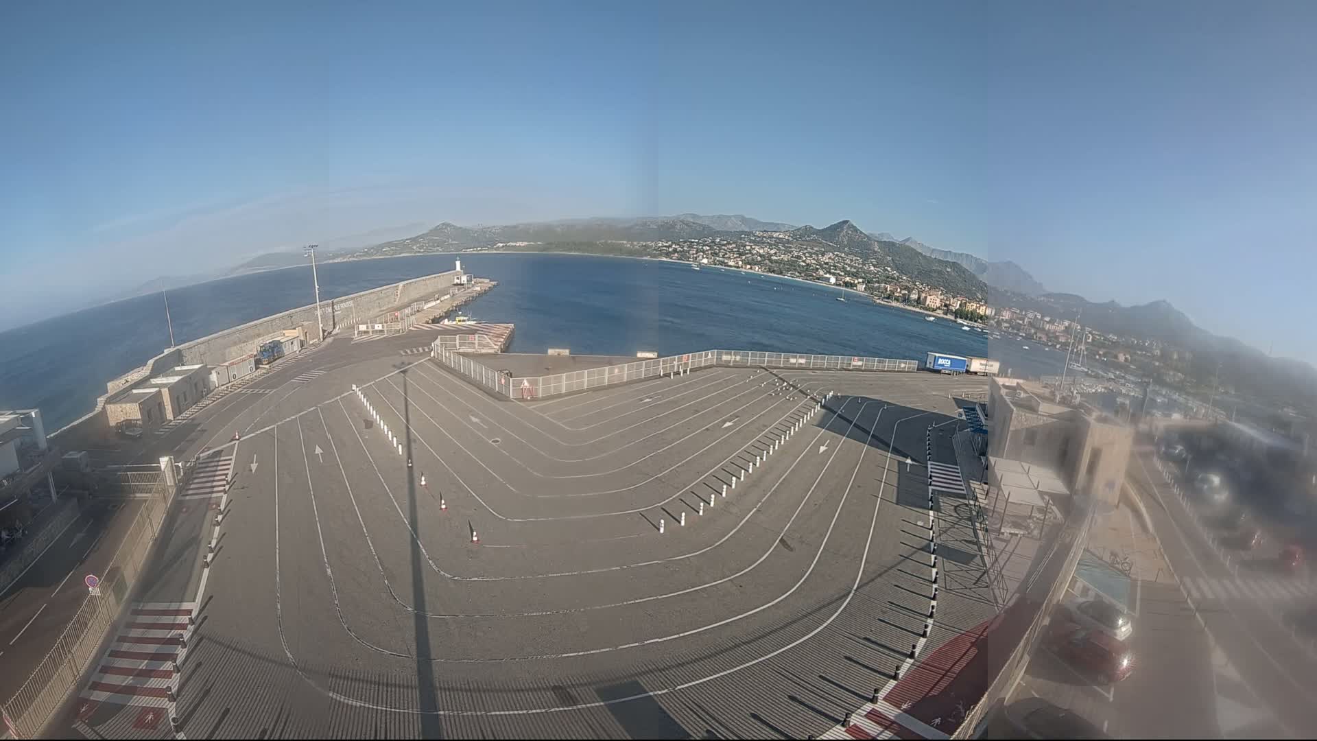 An empty, marked parking lot at a harbor overlooks a calm sea with distant mountains under a clear blue sky.