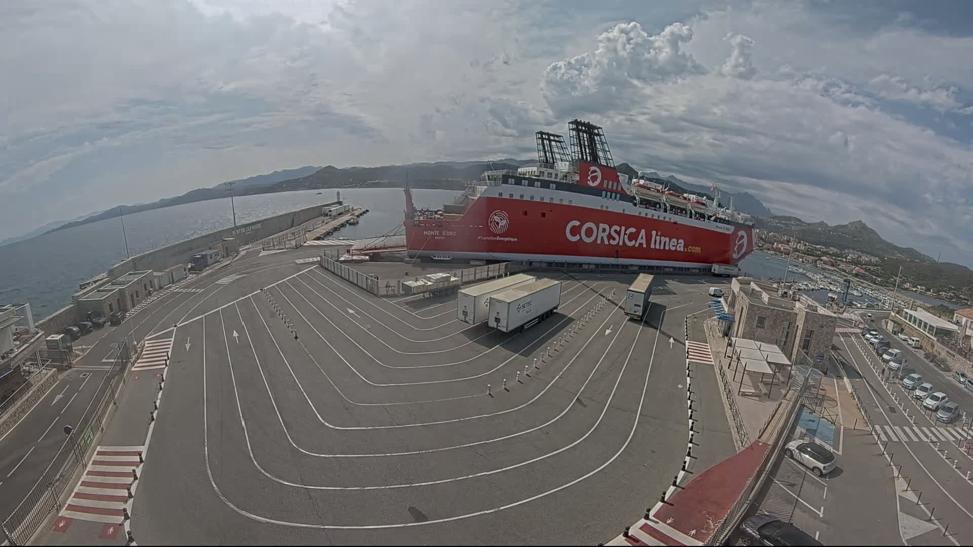A large red ferry is docked at a port under partly cloudy skies, with several vehicles parked nearby.