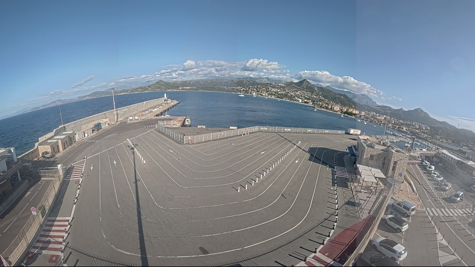 An empty ferry terminal parking lot with marked lanes and a few cars in an adjacent lot overlooks a calm bay with mountains in the background under a mostly clear sky.
