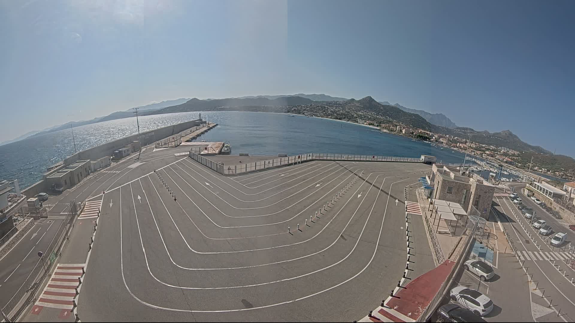 A ferry terminal with a large, curved parking lot overlooks a calm bay and mountains under a sunny sky.