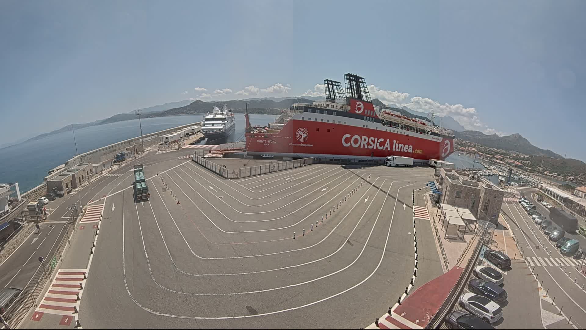A large red ferry is docked at a port on a sunny day with clear skies, alongside a smaller cruise ship, and several cars are parked nearby.