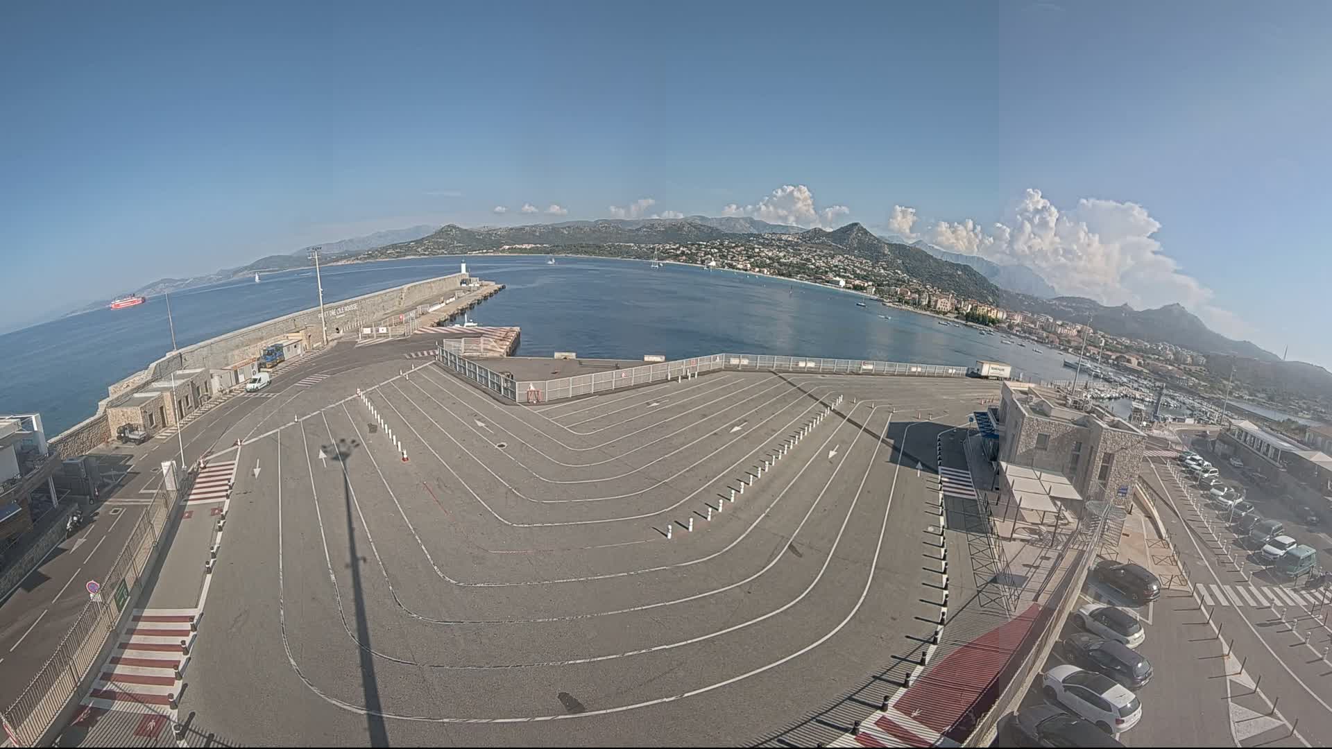 A large, empty vehicle staging area at a harbor, with a calm sea, mountains in the background, and partly cloudy skies.