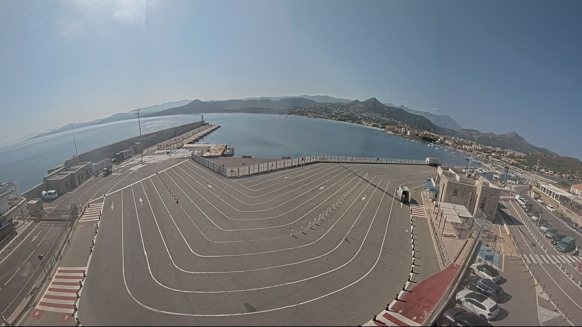 A harbor parking lot with marked lanes curves around a ferry terminal, overlooking calm blue water and distant mountains under a clear, sunny sky.