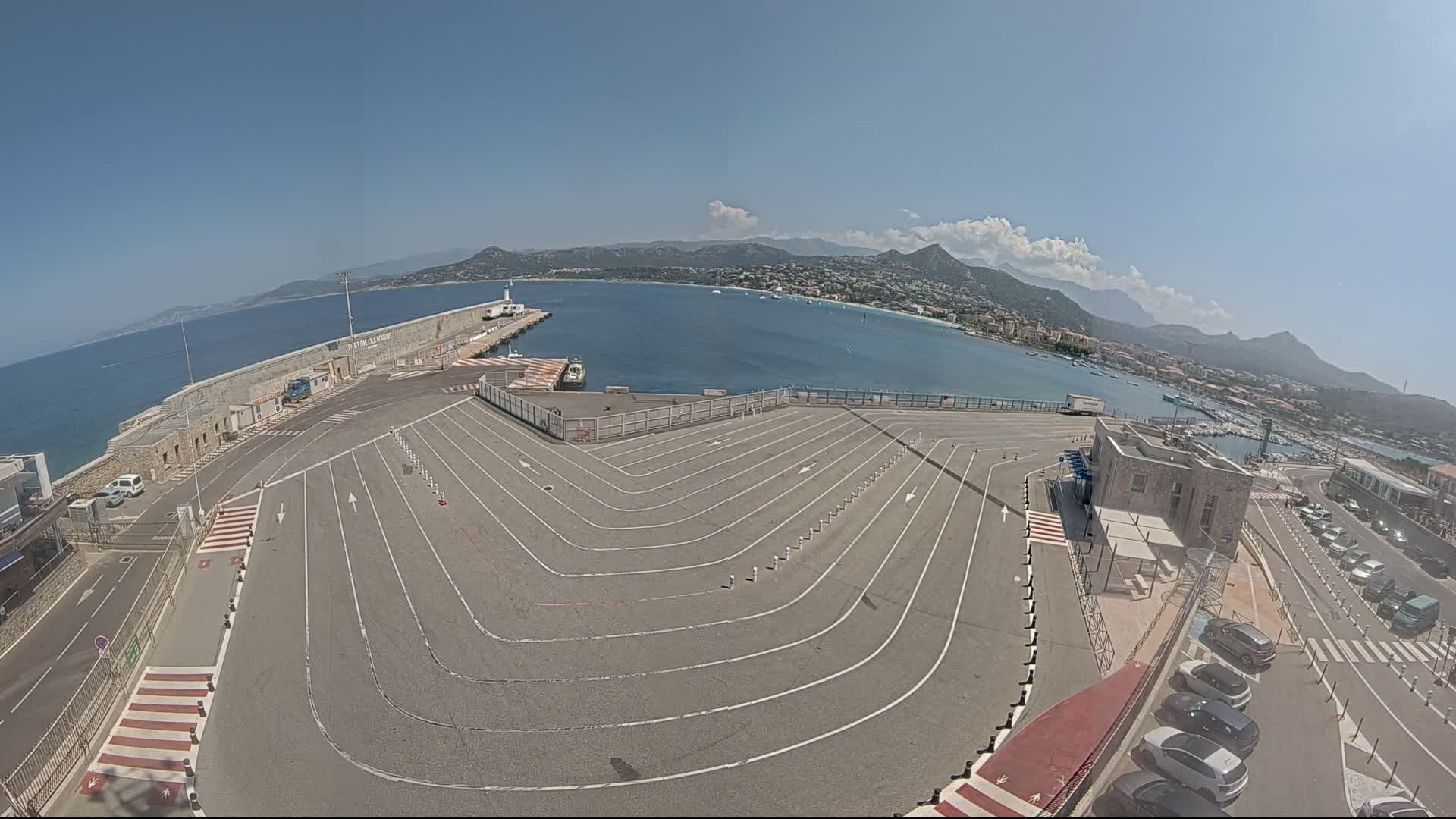 A large, empty asphalt area with painted lines, possibly a ferry terminal, overlooks a calm bay with mountains in the background under a clear, sunny sky.