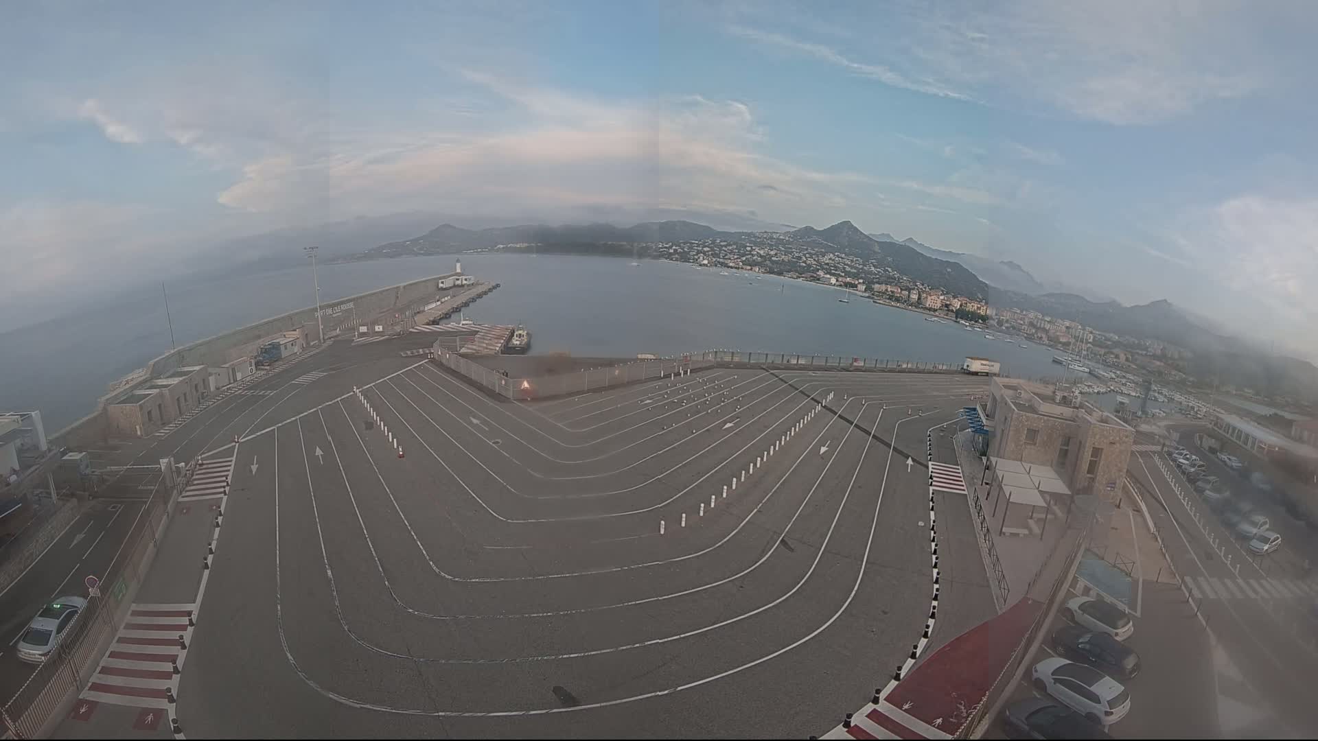 A mostly empty, curved parking lot overlooks a calm harbor with distant mountains under a hazy sky.