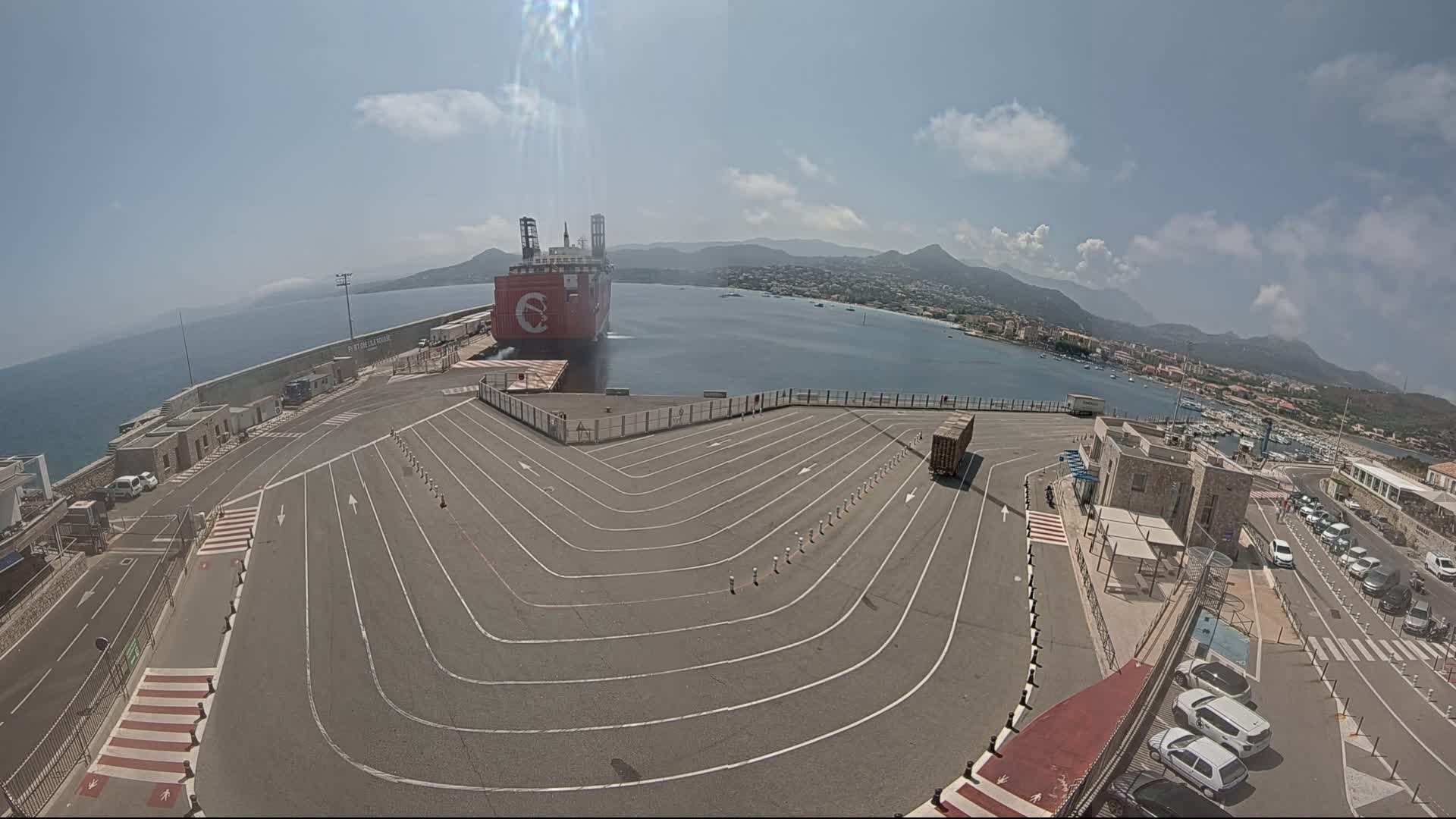 A large red cargo ship is docked at a port on a sunny day, with a paved area marked with driving lanes and several cars parked nearby, and mountains visible in the distance.
