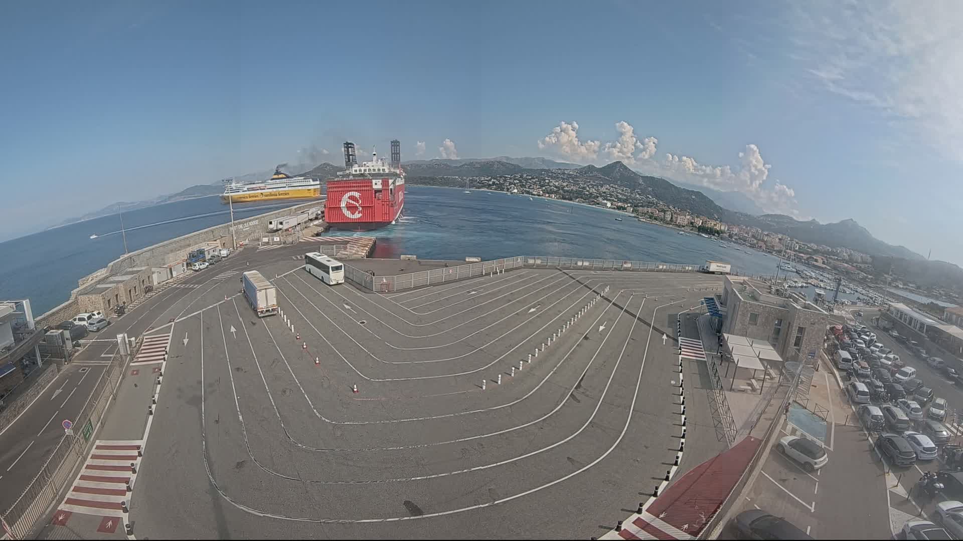 A large red and white ferry is docked at a port on a sunny day with partly cloudy skies,  and a paved area with marked lanes for vehicles is visible, along with parked cars and a bus.