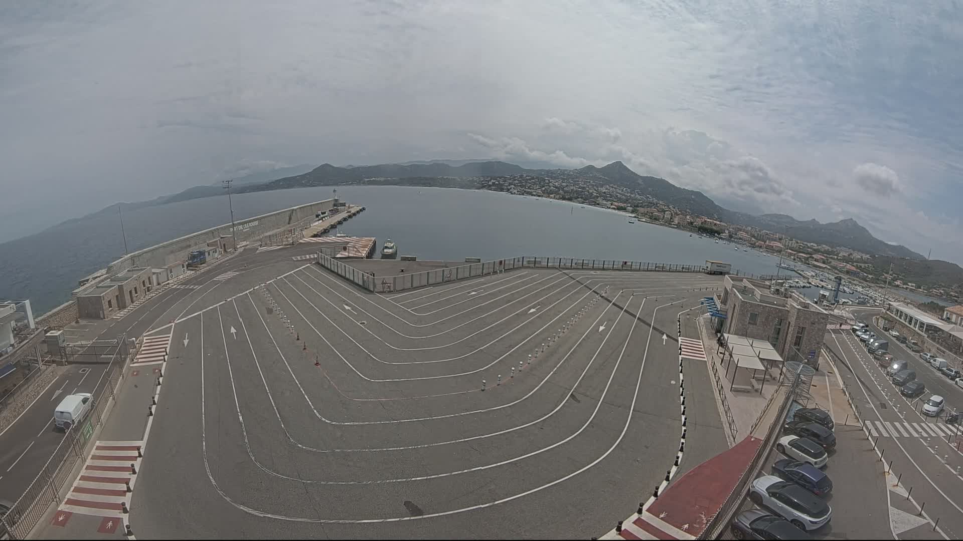 An aerial view shows a ferry terminal with a large, curving vehicle queueing area next to a calm body of water under a partly cloudy sky.