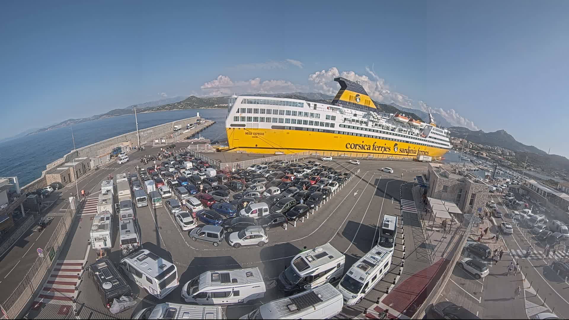 A large yellow ferry is docked at a port on a sunny day, surrounded by a large parking lot filled with cars and RVs.