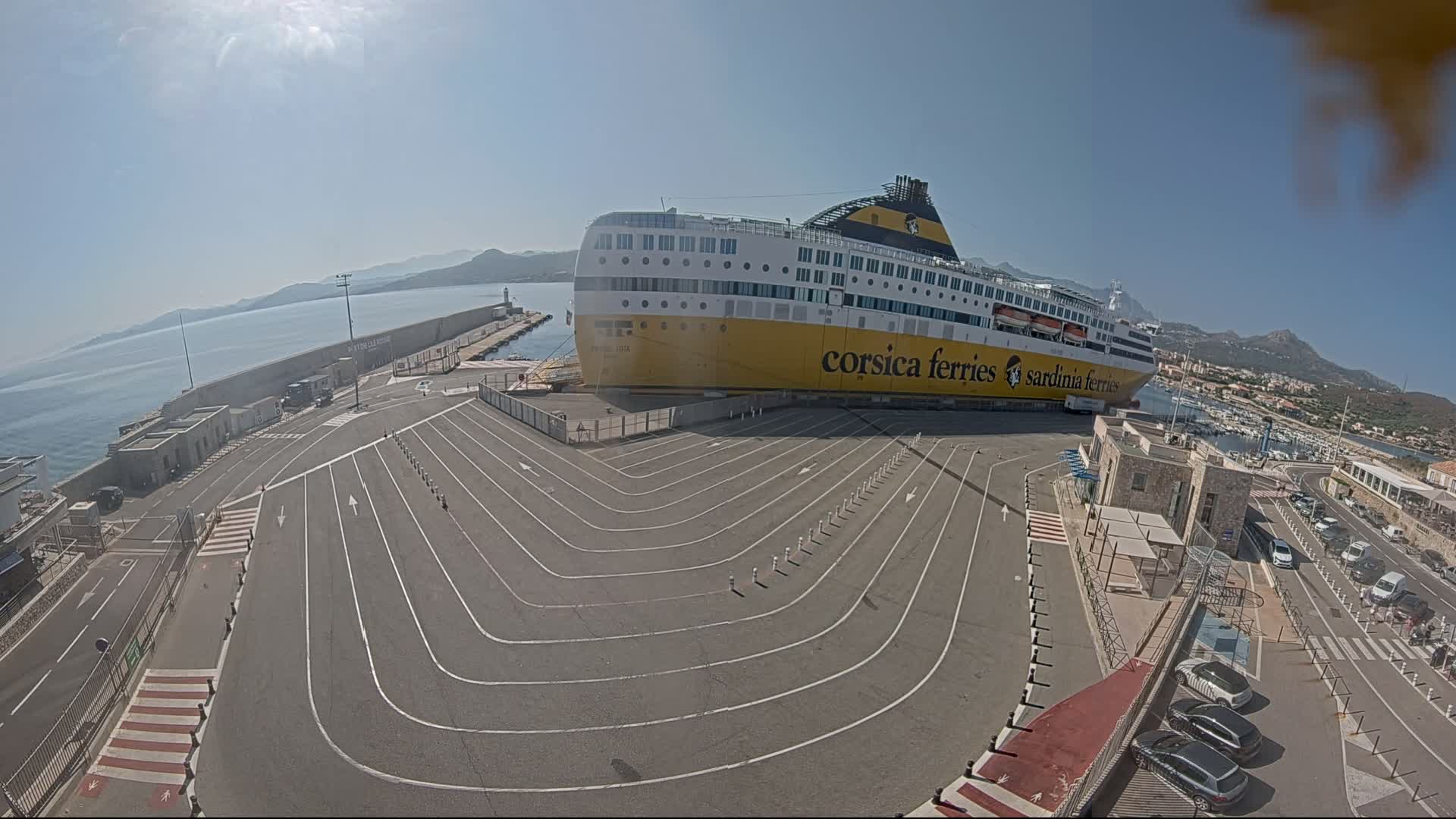 A large ferry is docked at a port on a sunny day, with a paved area marked with lines for vehicles leading to it.