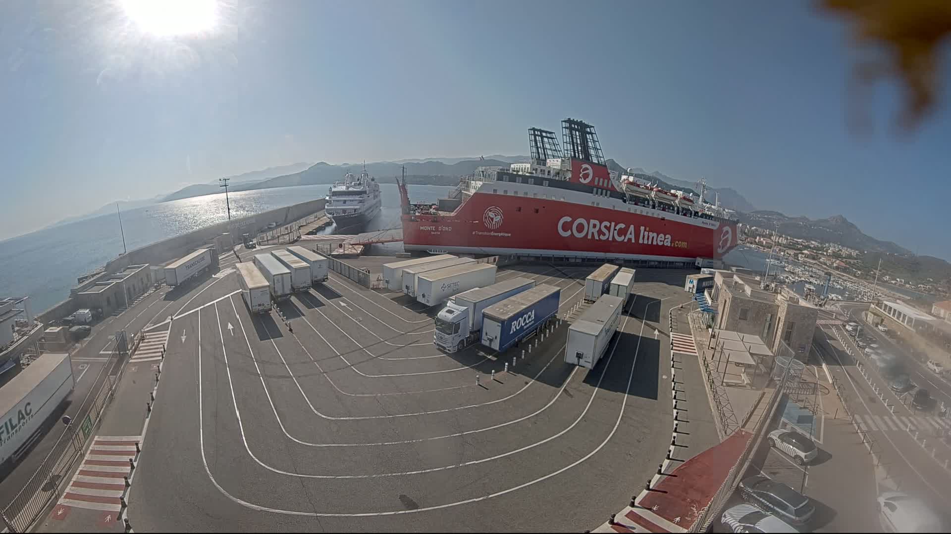 A large red ferry is docked at a port on a sunny day, with several cargo trucks parked nearby and another smaller ship visible in the distance.