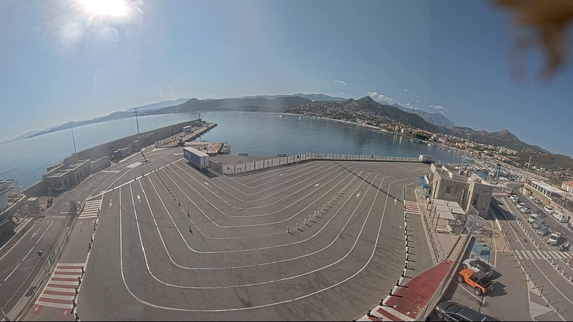 A sunny day at a harbor shows a large, empty, lined parking area next to a calm body of water with mountains in the background.