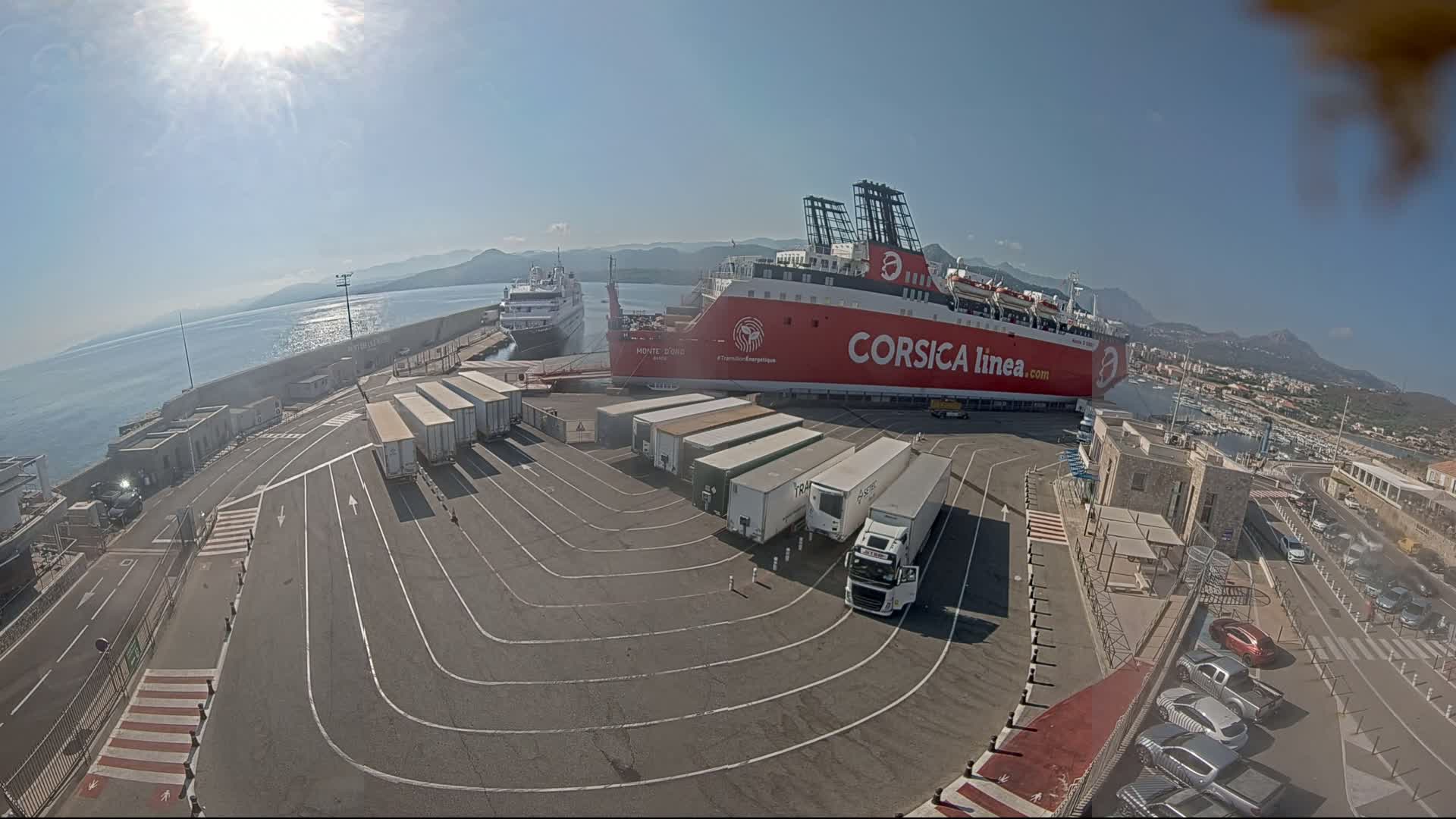 A large red ferry is docked at a port on a sunny day, surrounded by trucks and cars, with another smaller ship visible in the background.