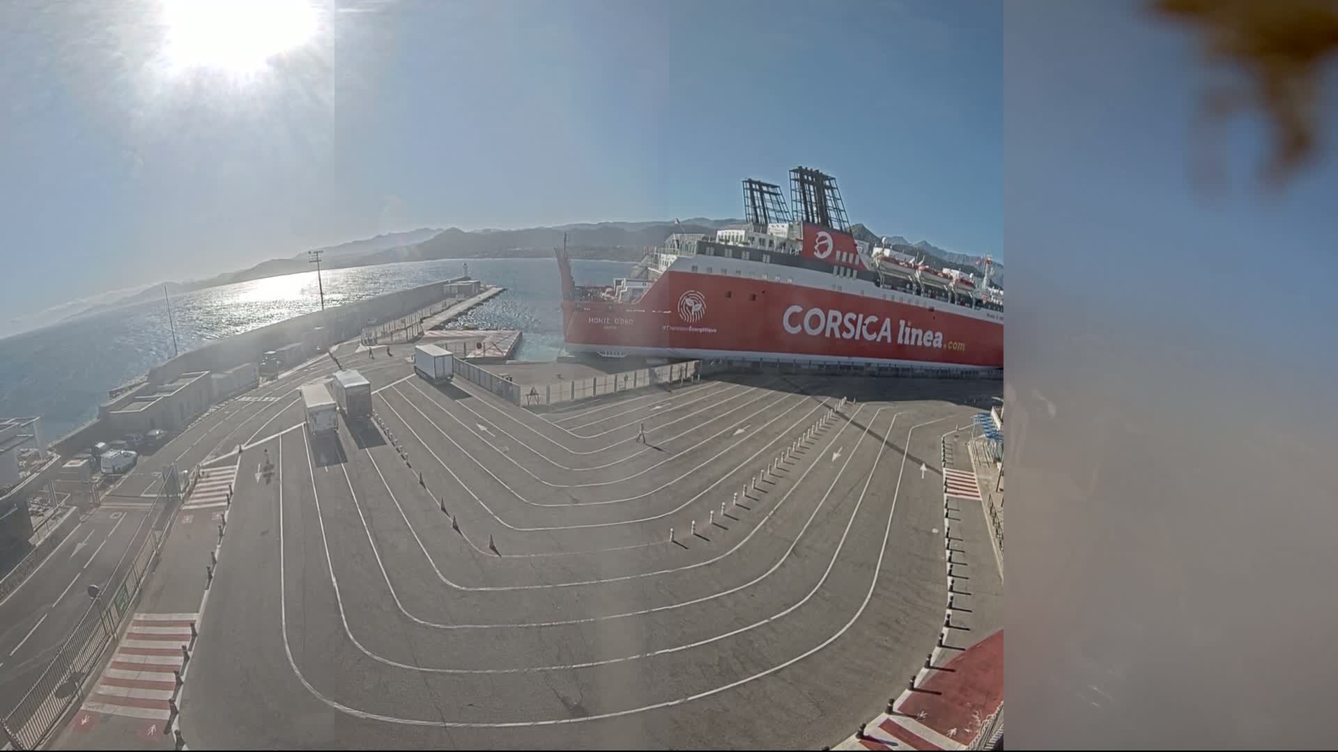 A large red and white ferry is docked at a port on a sunny day, with several vehicles parked in designated lanes on the dock.