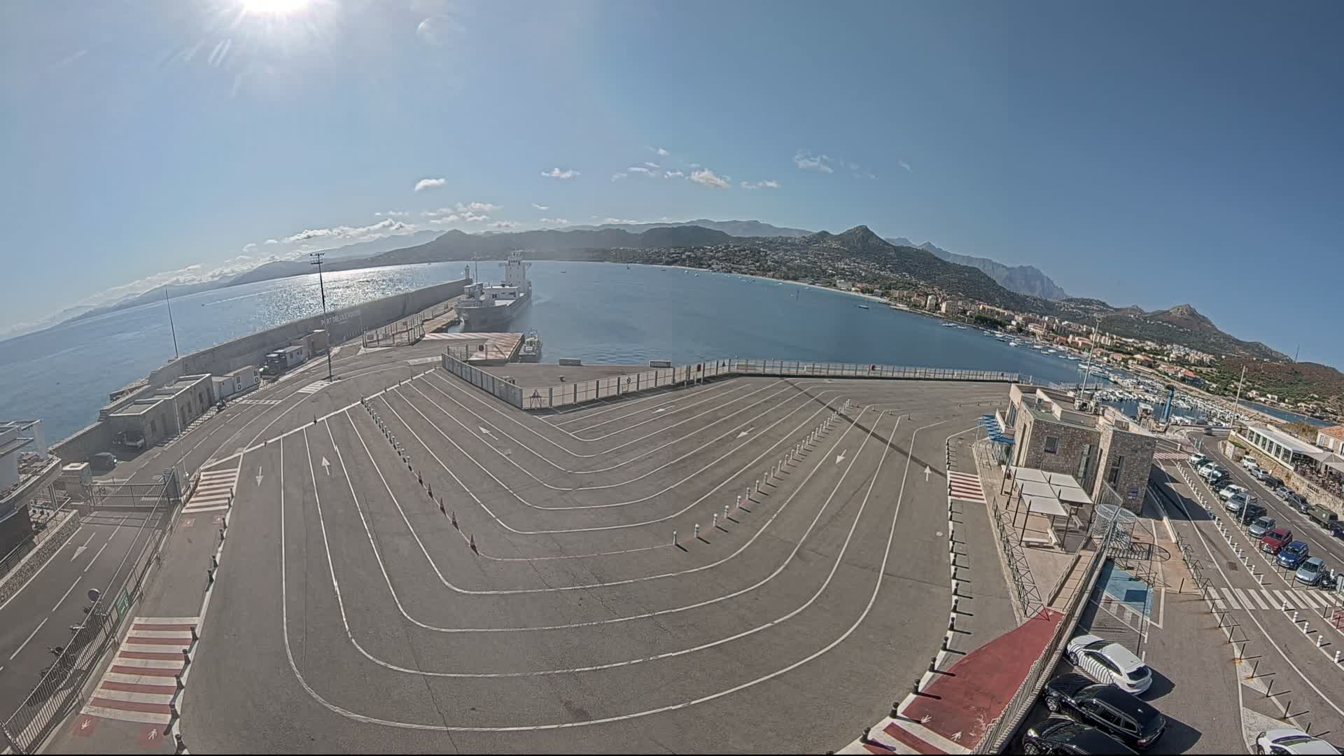 A sunny harbor scene shows a large, empty vehicle staging area next to a body of water with a town and mountains in the background.
