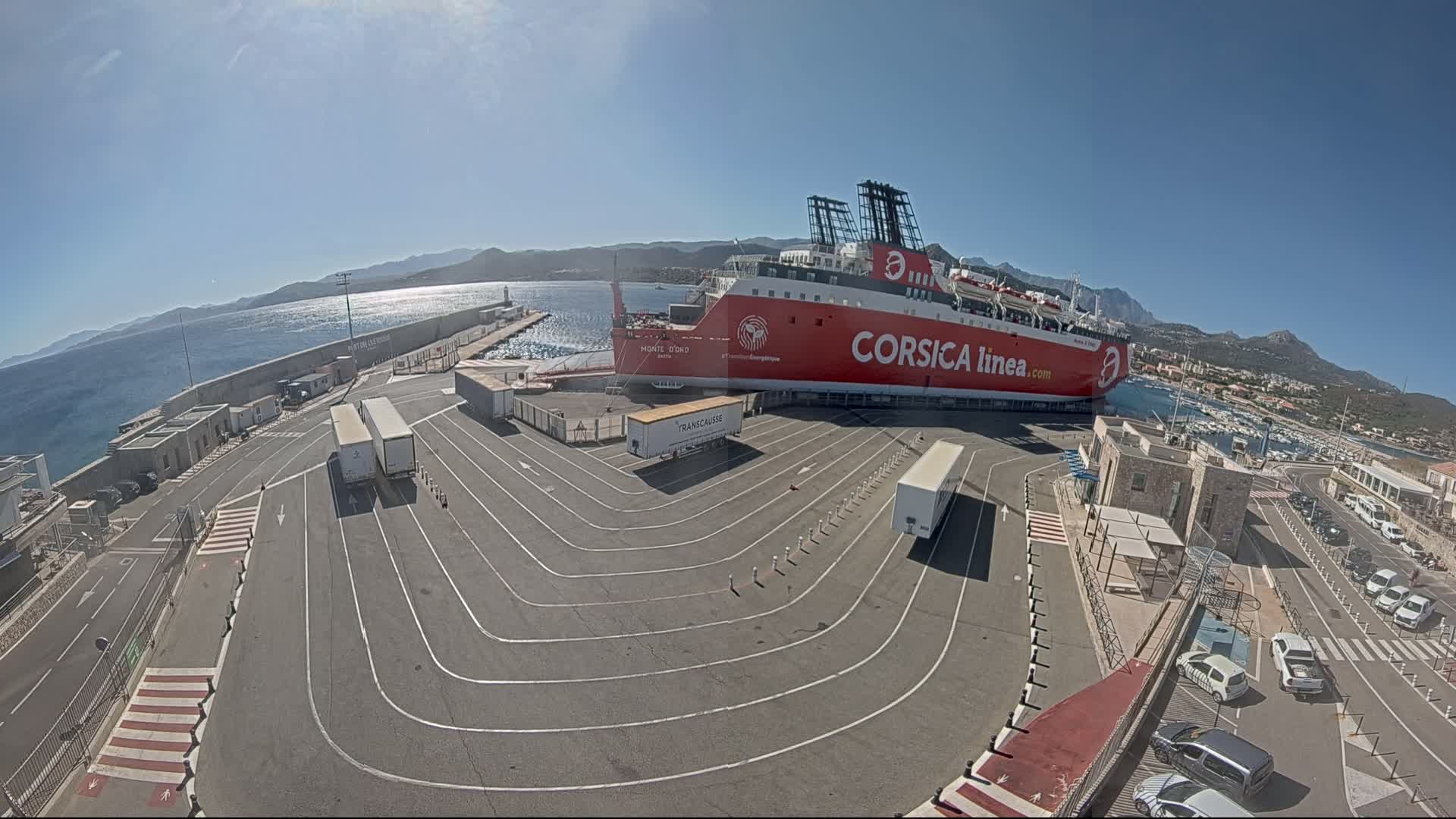 A large red ferry is docked at a port on a sunny day, with several trucks and cars parked in designated lanes nearby.