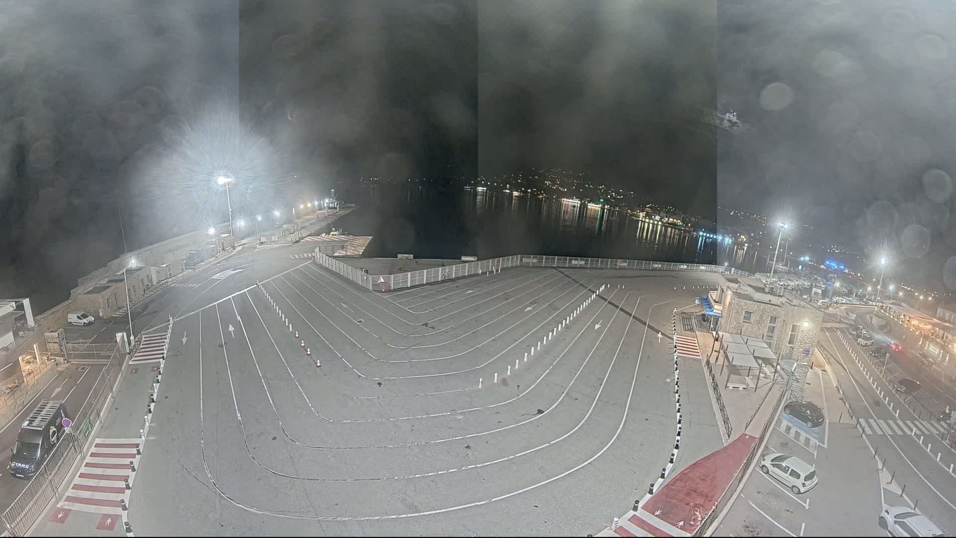 A nighttime, hazy view of a port area shows a large, empty vehicle staging area with lane markings next to a body of water where lights from a town are reflected.
