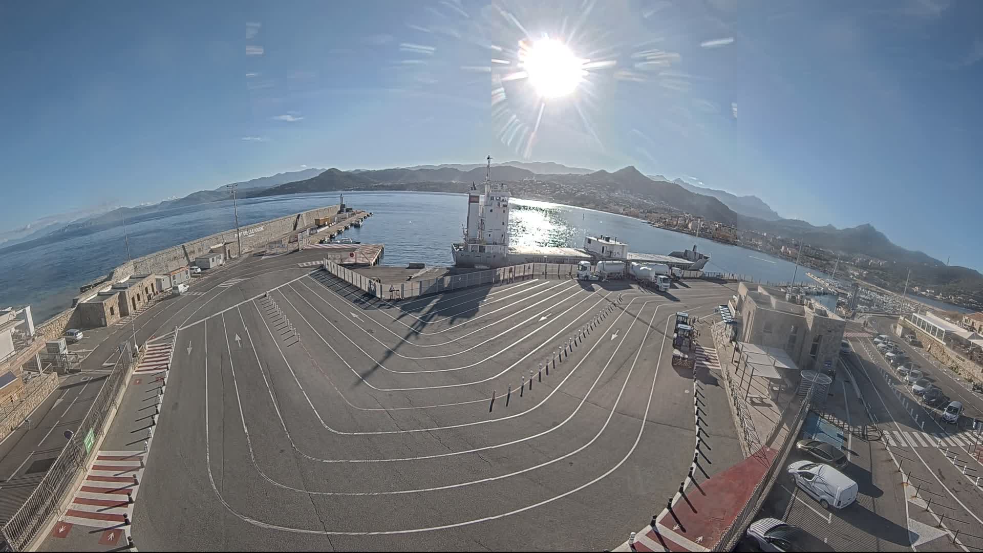 Under a radiant sun in a clear blue sky, a wide-angle view reveals a bustling port scene with a ship docked at a concrete pier, a large paved vehicle staging area, and a coastal town nestled against distant mountains.