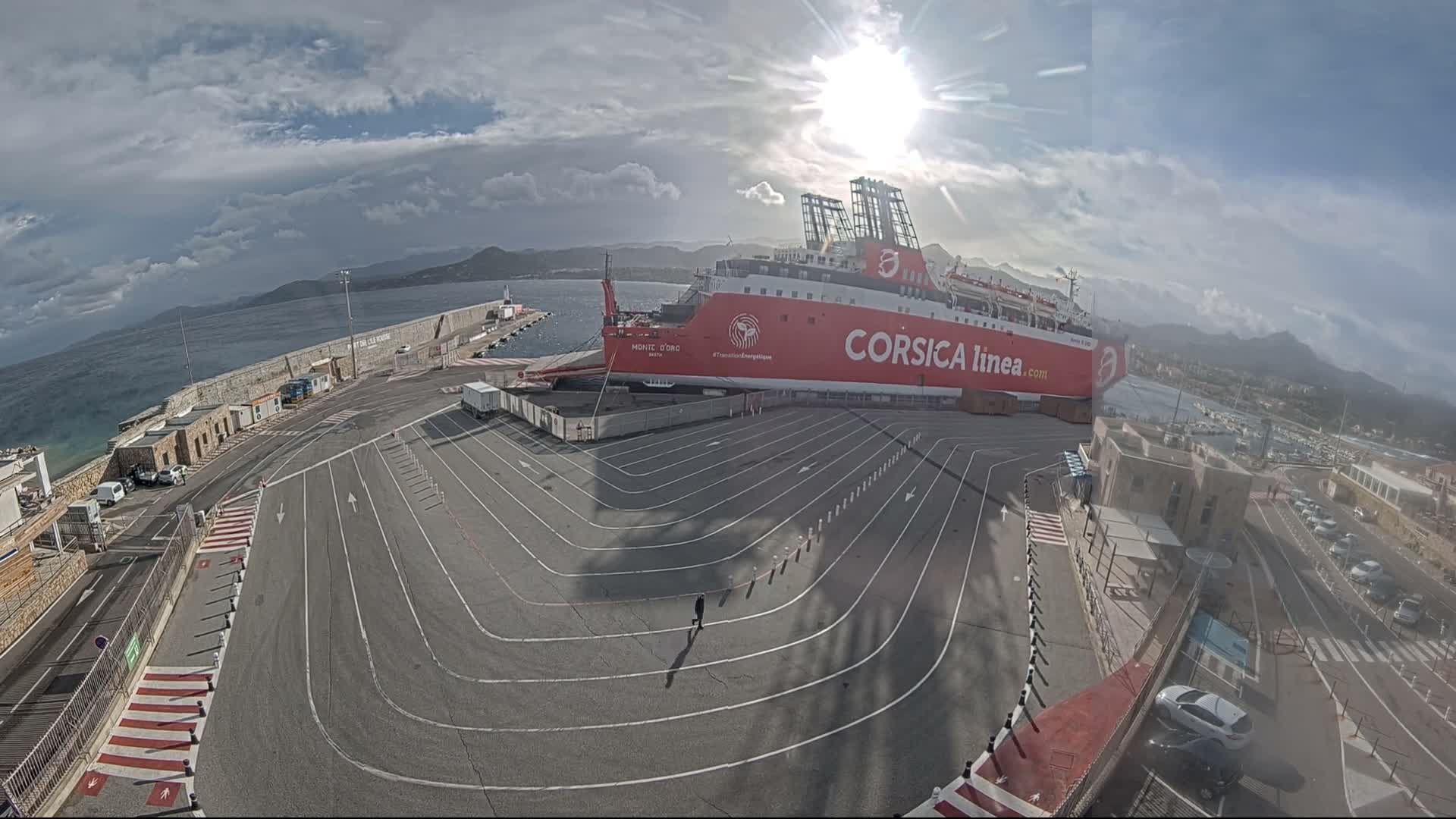 A wide-angle view captures a large red ferry docked at a port with a spacious, marked ground and a lone person walking, all under a partly cloudy sky with brilliant sunshine illuminating distant mountains.