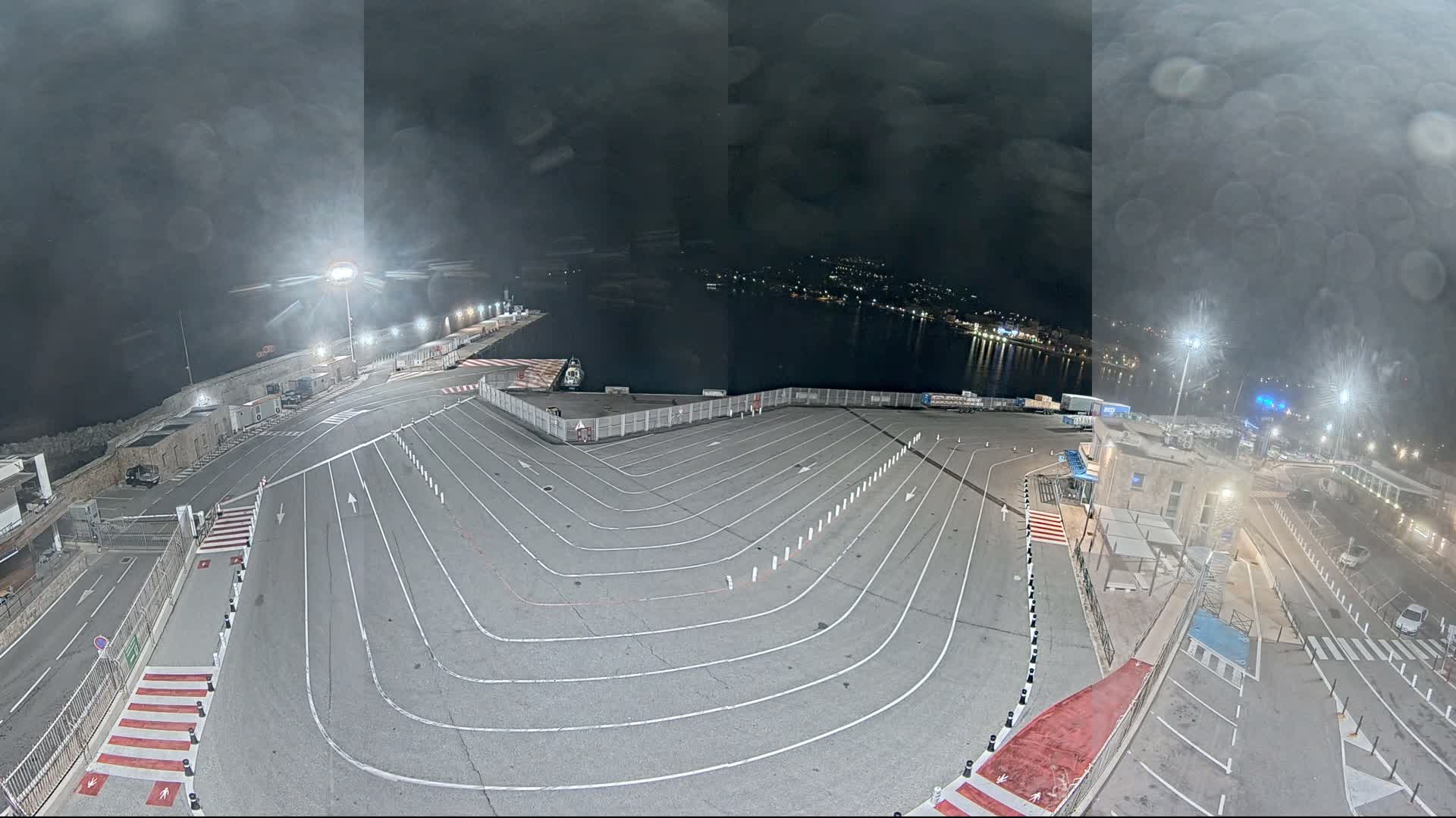 A wide-angle view captures a large red ferry docked at a port with a spacious, marked ground and a lone person walking, all under a partly cloudy sky with brilliant sunshine illuminating distant mountains.