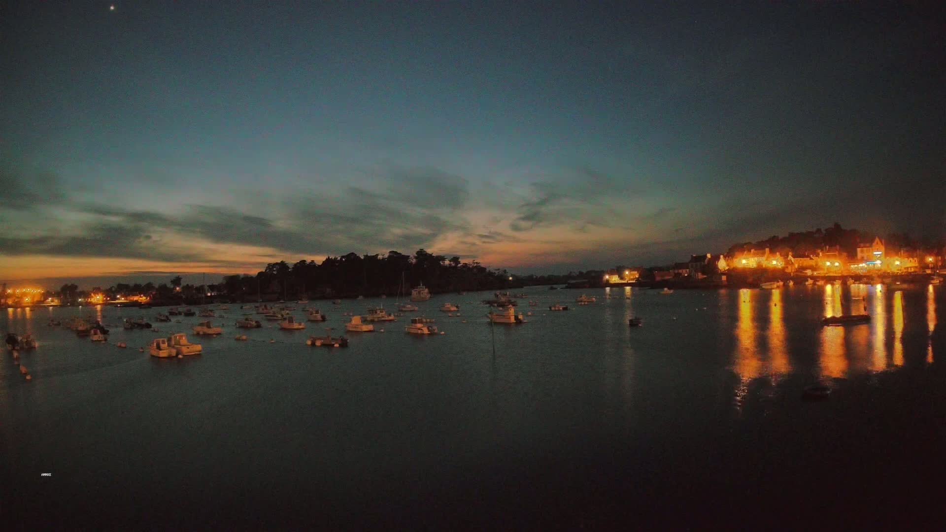 Many small boats are moored in a calm harbor at twilight, with a town's lights reflecting on the water.