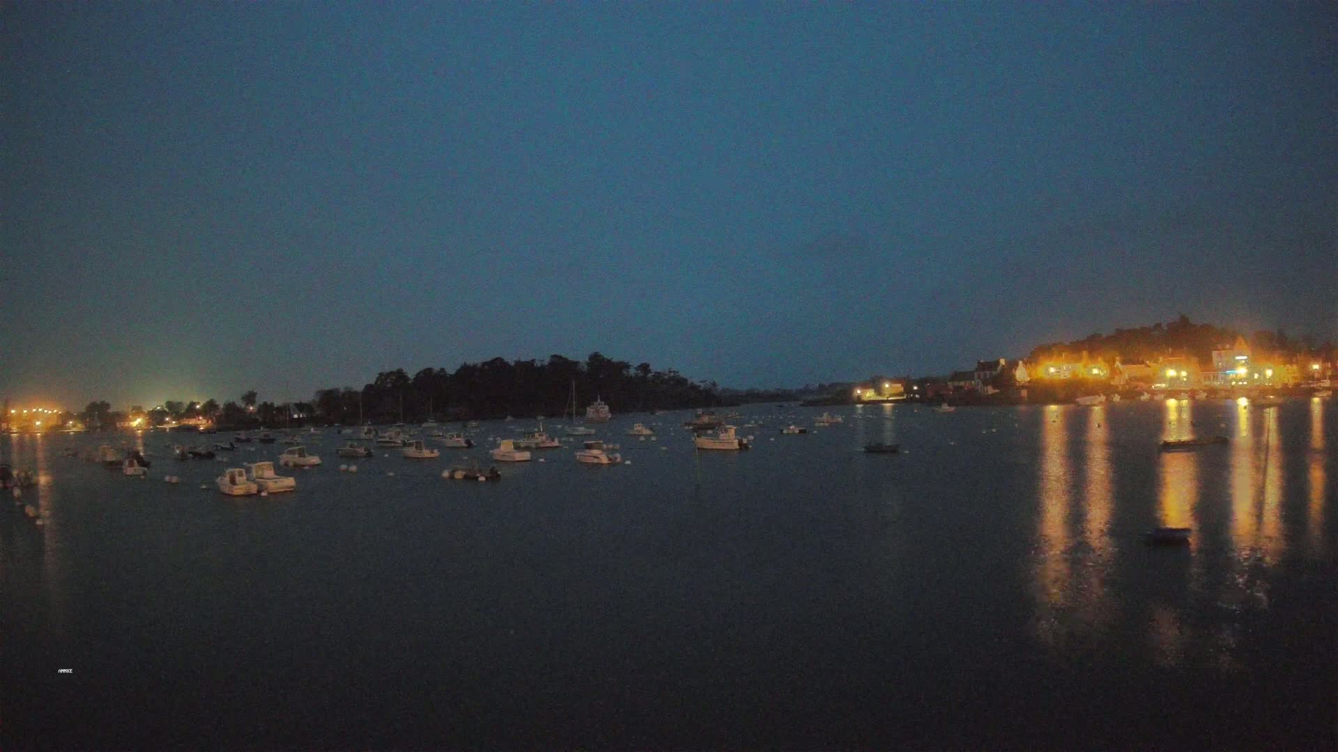 Many small boats are moored in a calm, dark-blue body of water at night, with the lights of a town reflecting on the water in the distance.