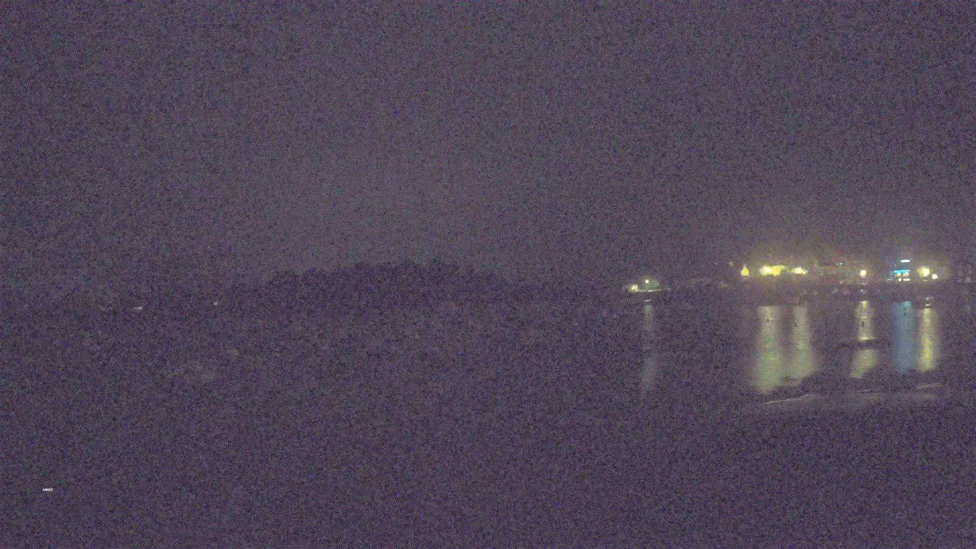 A nighttime view of a dark shoreline with faintly lit buildings and calm water reflecting the lights.