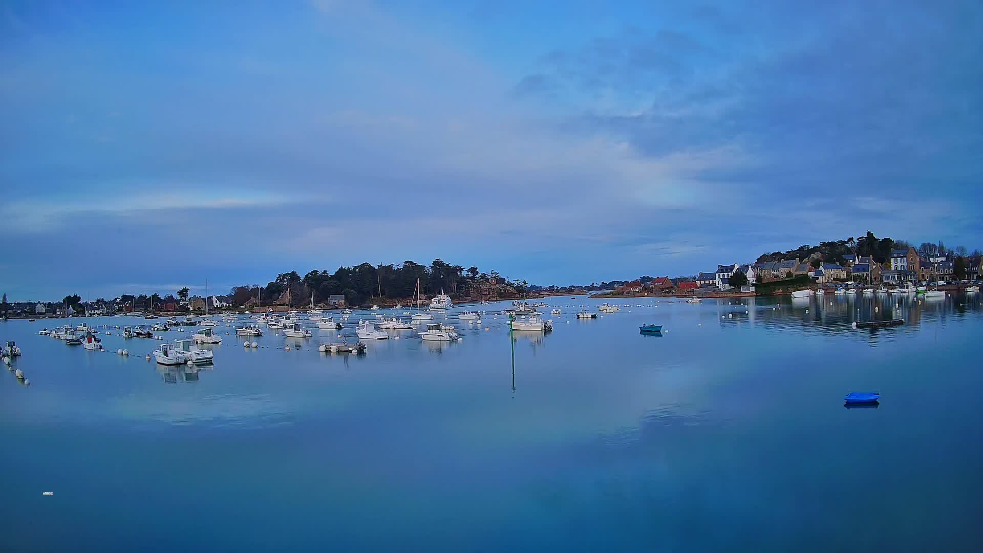 Numerous white boats are moored in a calm, blue bay under a mostly clear, light-blue sky, with a small village visible on the far shore.