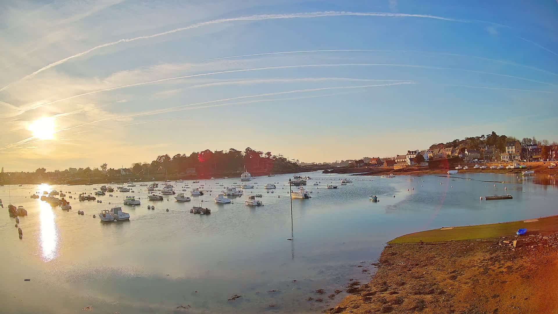 Many small boats are moored in a calm harbor at sunset under a clear sky with contrails.