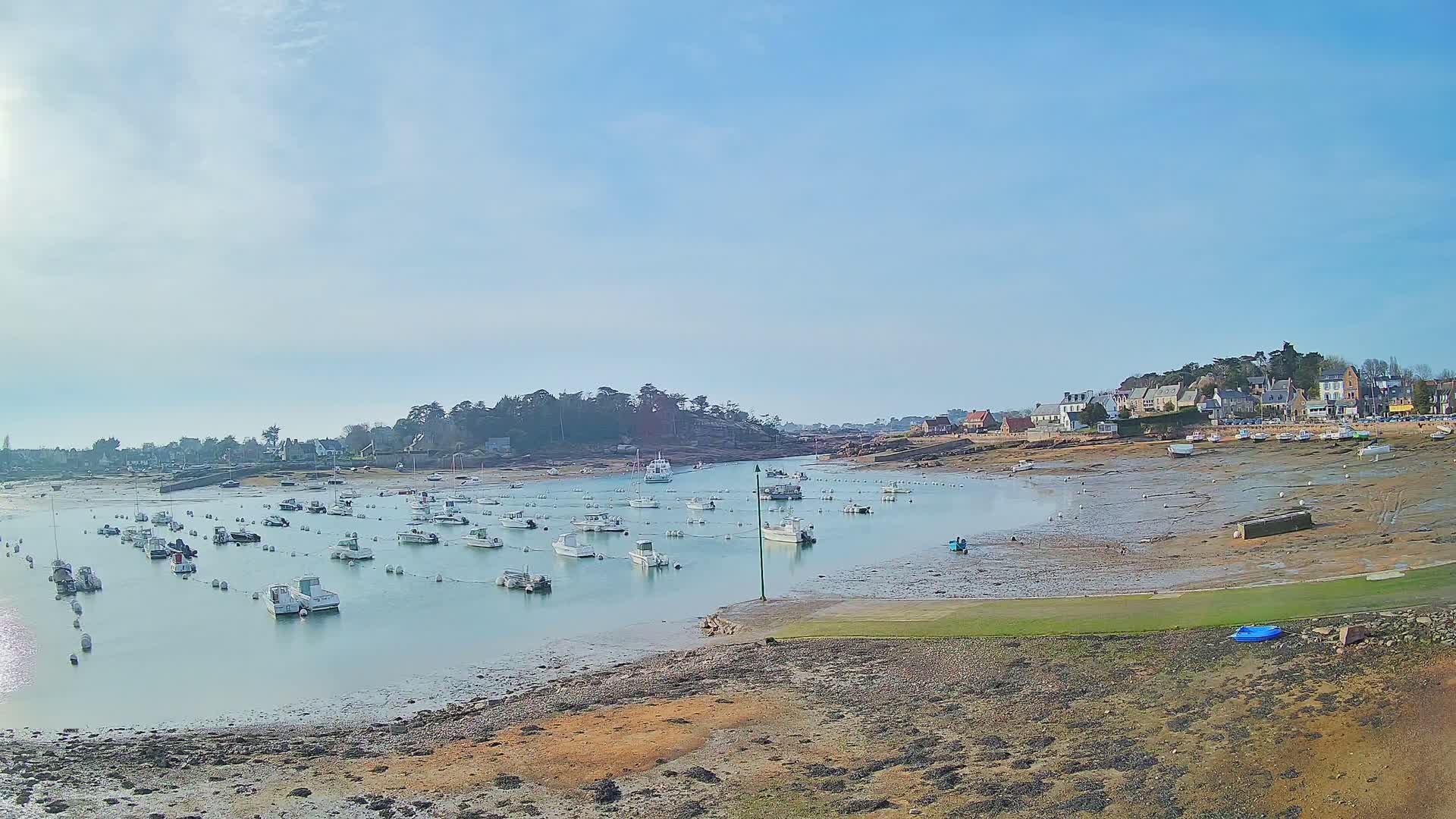 Under a mostly clear sky, numerous small white boats are moored in a calm harbor at low tide, with a small village visible in the background.