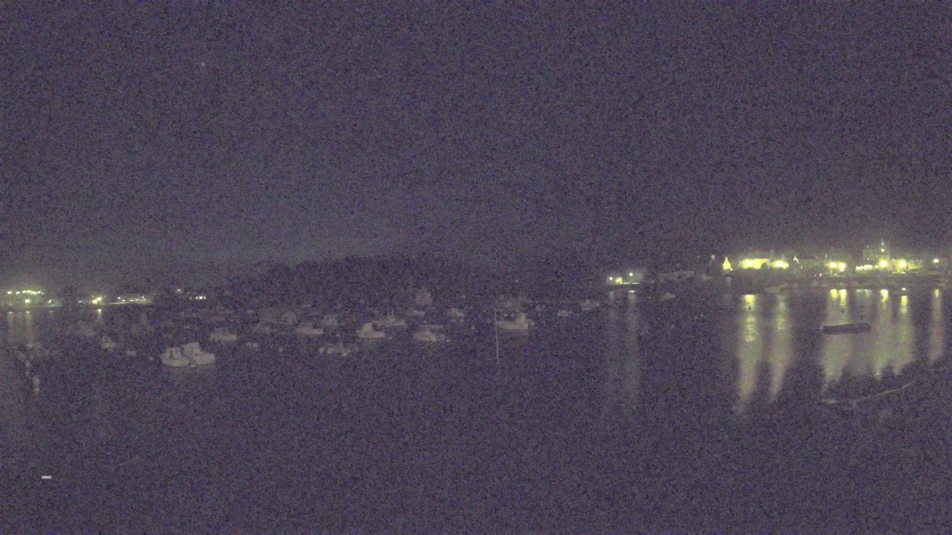 A nighttime view of a harbor filled with boats, with a town's lights reflecting on the calm water in the background.