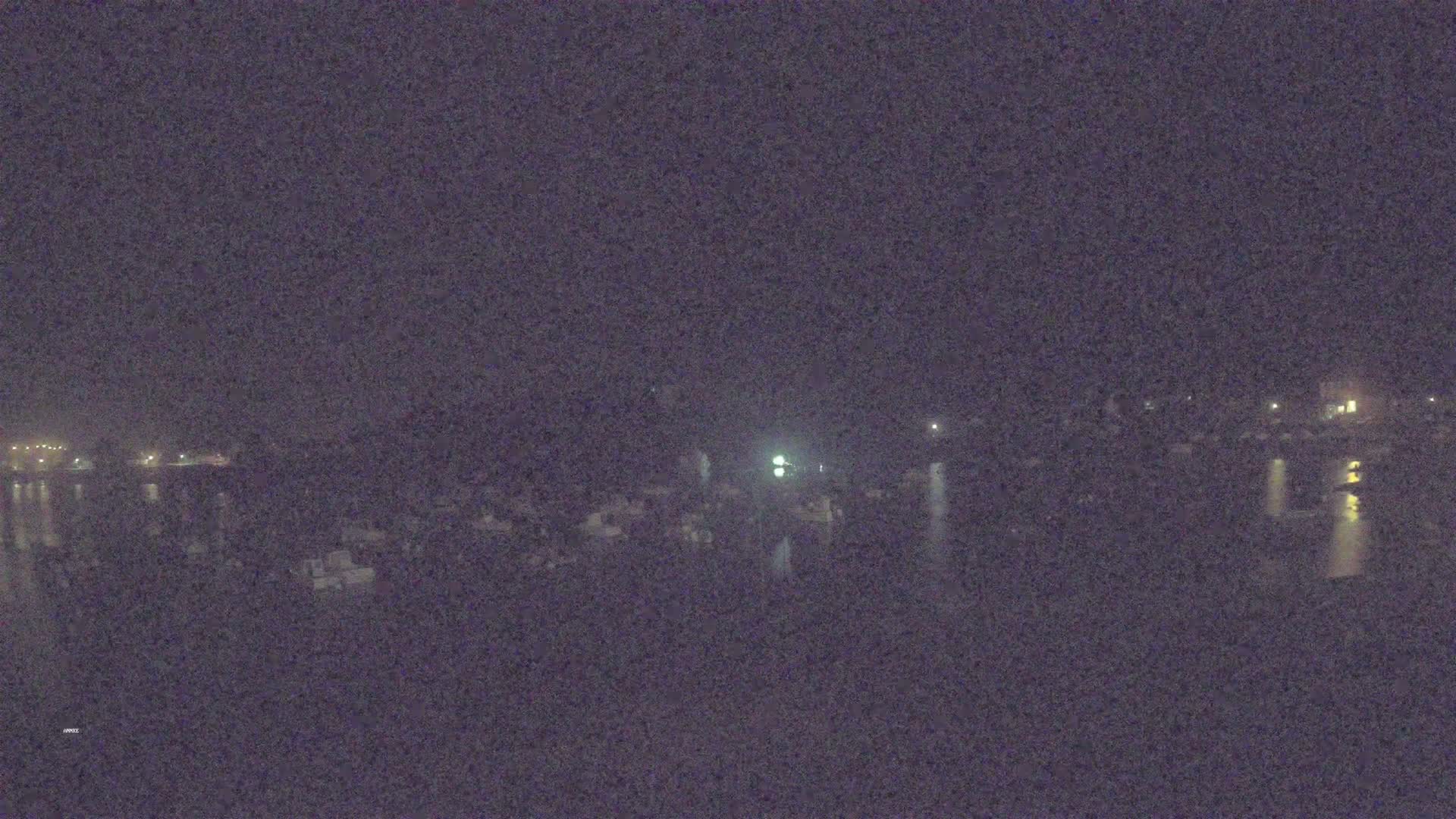 A nighttime, low-light view shows a body of water reflecting scattered lights from a shoreline with buildings and vehicles.