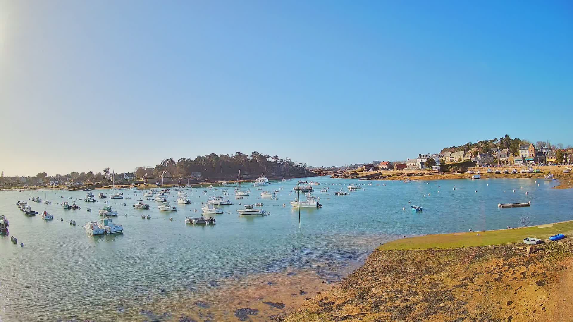 Many small white boats are moored in a calm, shallow bay under a clear blue sky, with a small village visible on the far shore.