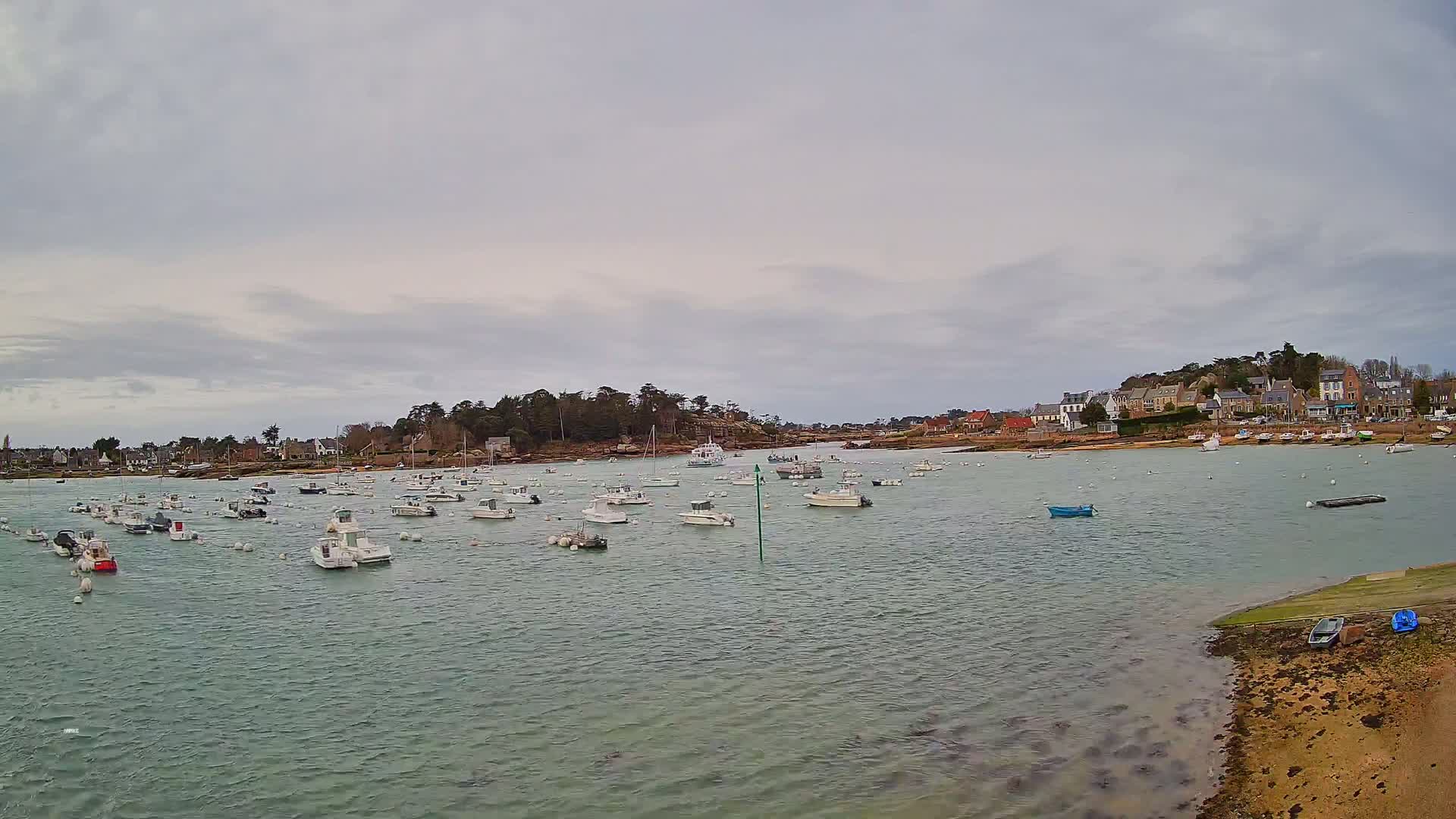 Numerous small boats are moored in a calm bay under an overcast sky, with a small village visible on the far shore.