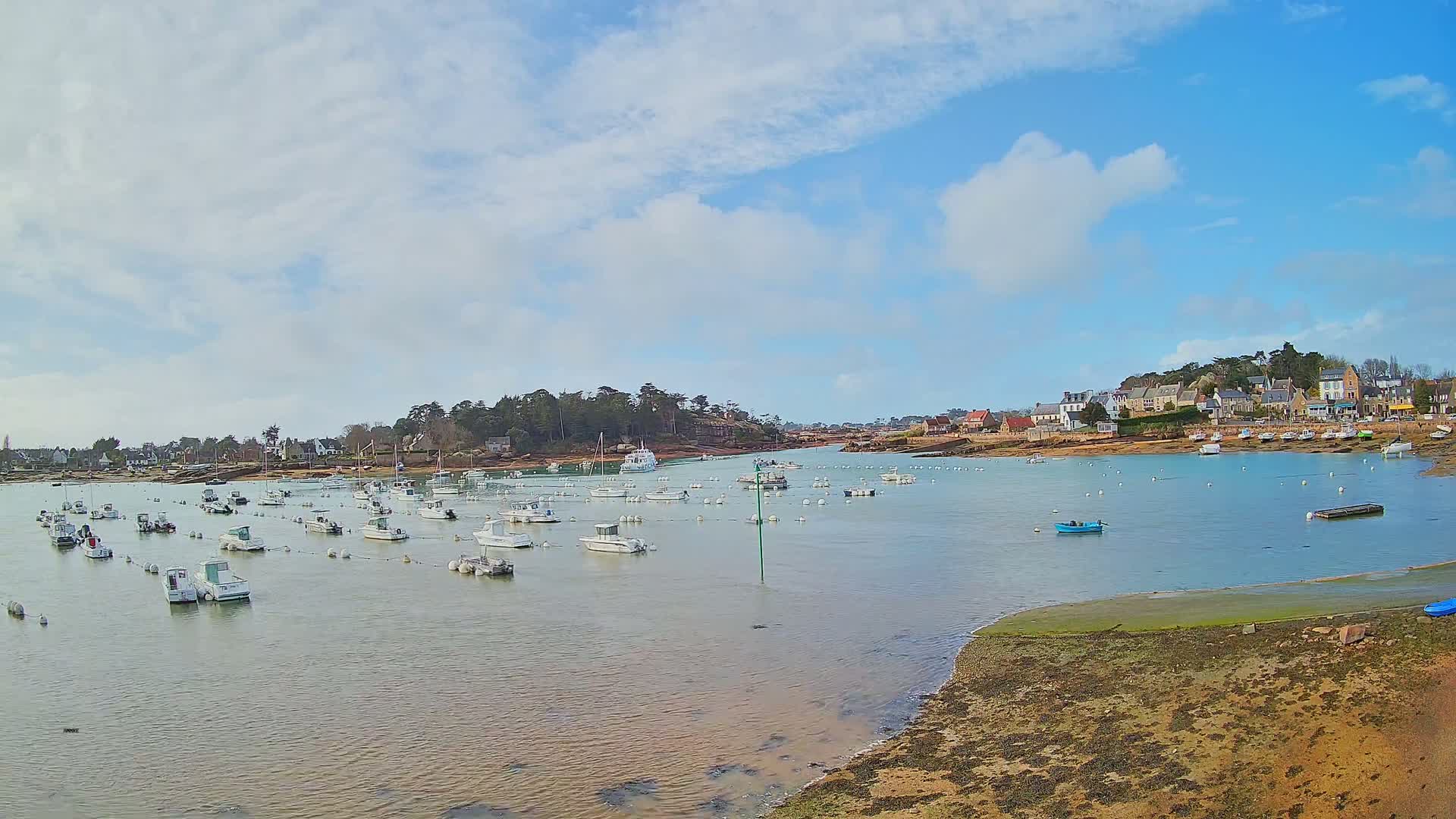 A calm bay with numerous small boats moored, a small village nestled on the shore under a partly cloudy sky.