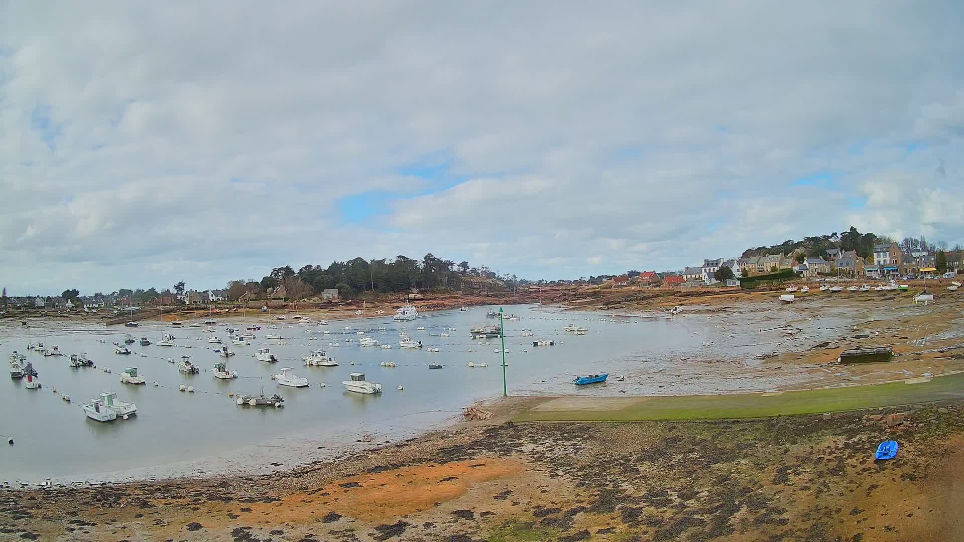 Many small boats are moored in a calm bay under a cloudy sky, with a small village visible in the background and exposed mudflats at low tide.