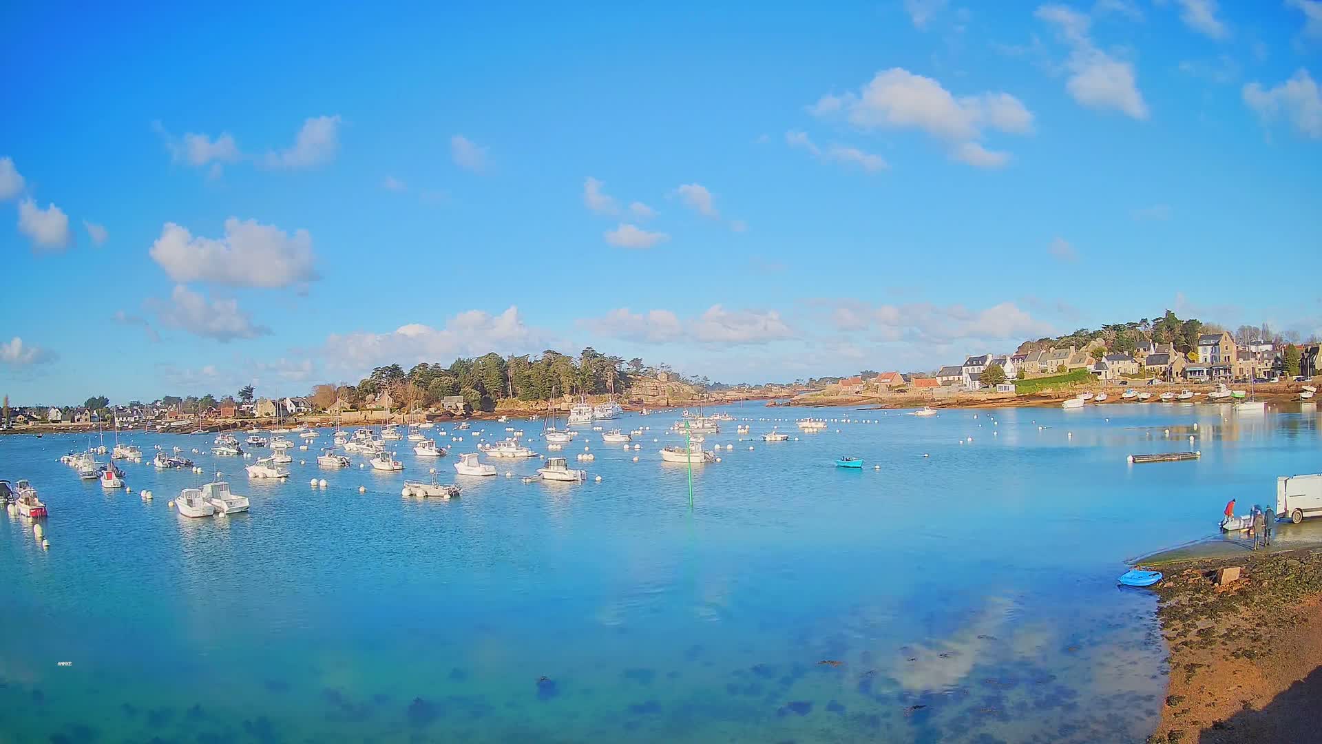 Under a bright, sunny sky with a few scattered clouds, a calm, turquoise bay is filled with numerous small white boats moored near a shoreline village.