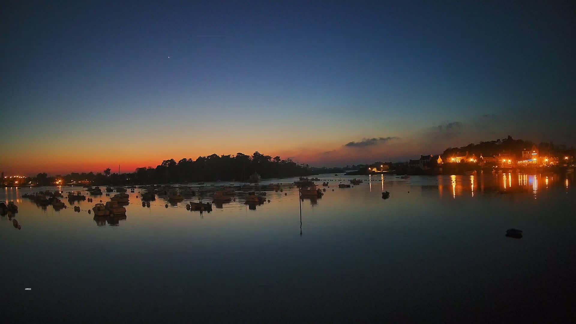 Many small boats are moored in a calm body of water at dusk, under a twilight sky, with a village’s lights reflecting in the water.