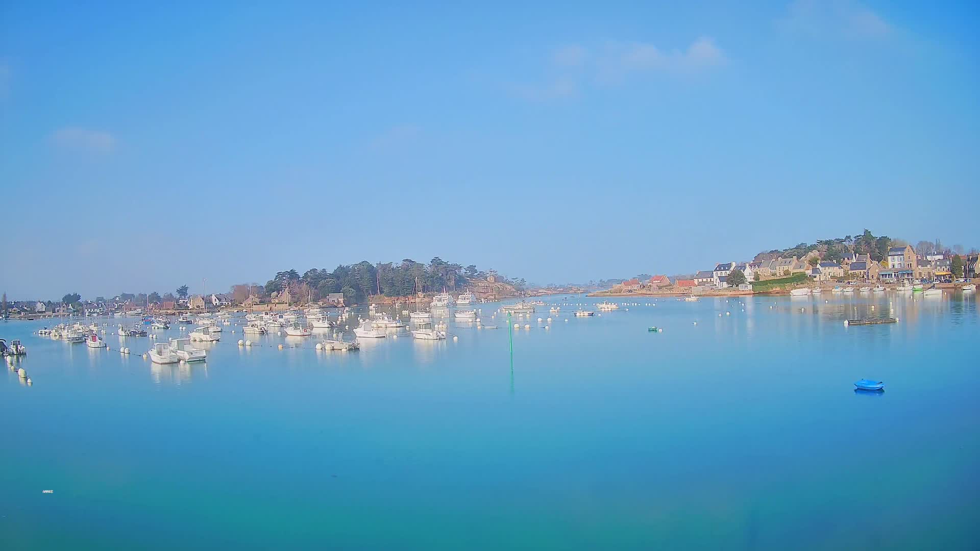Numerous white boats are moored in a calm, turquoise harbor, with a small village and a tree-lined peninsula visible in the background under a clear blue sky.