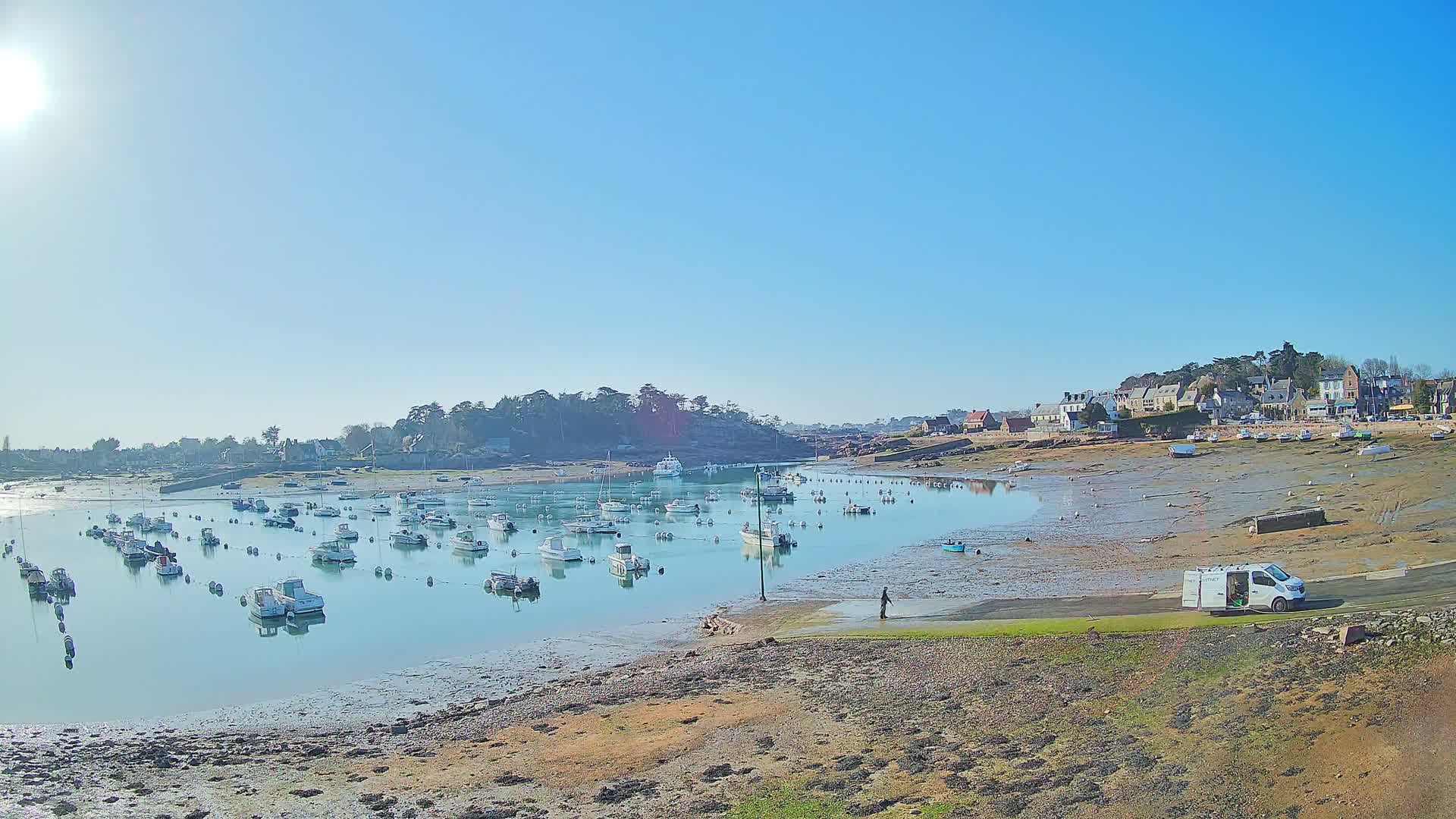 Under a clear, sunny sky, numerous small boats are moored in a calm harbor at low tide, with a small village and a van visible in the background.